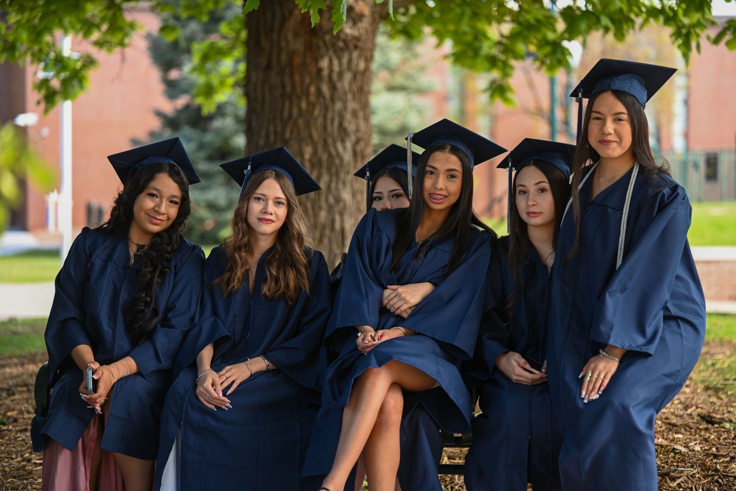 Six female graduates in navy blue caps and gowns pose together outdoors near a tree on a college campus.