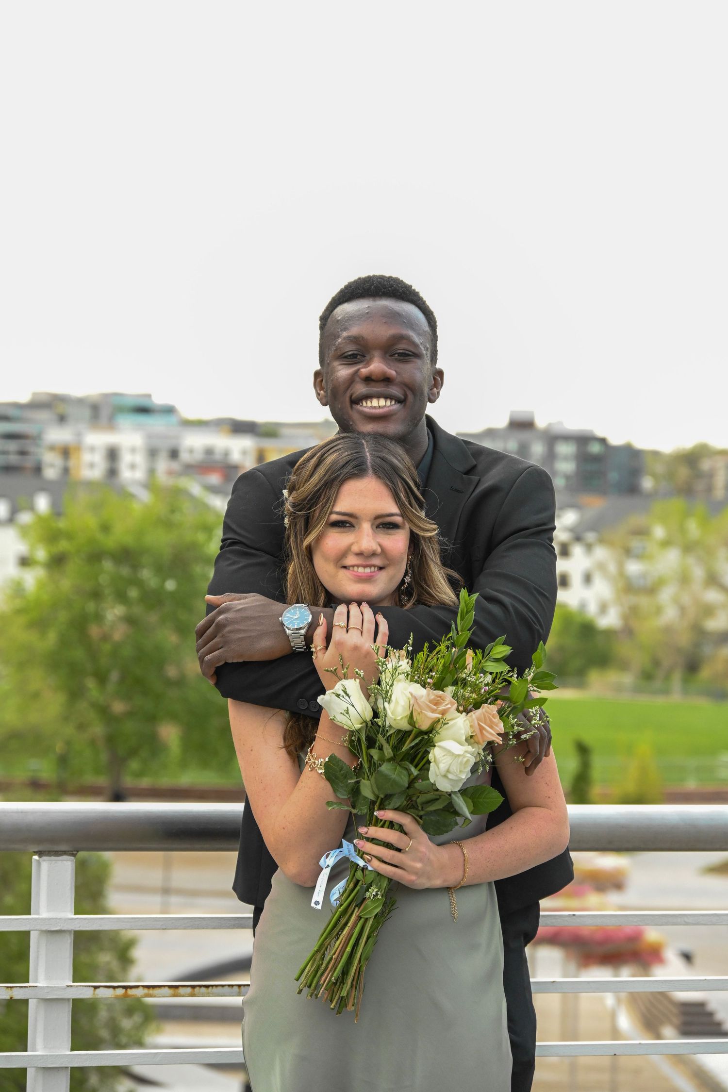 Smiling couple poses on bridge, man embracing woman holding bouquet of white roses.