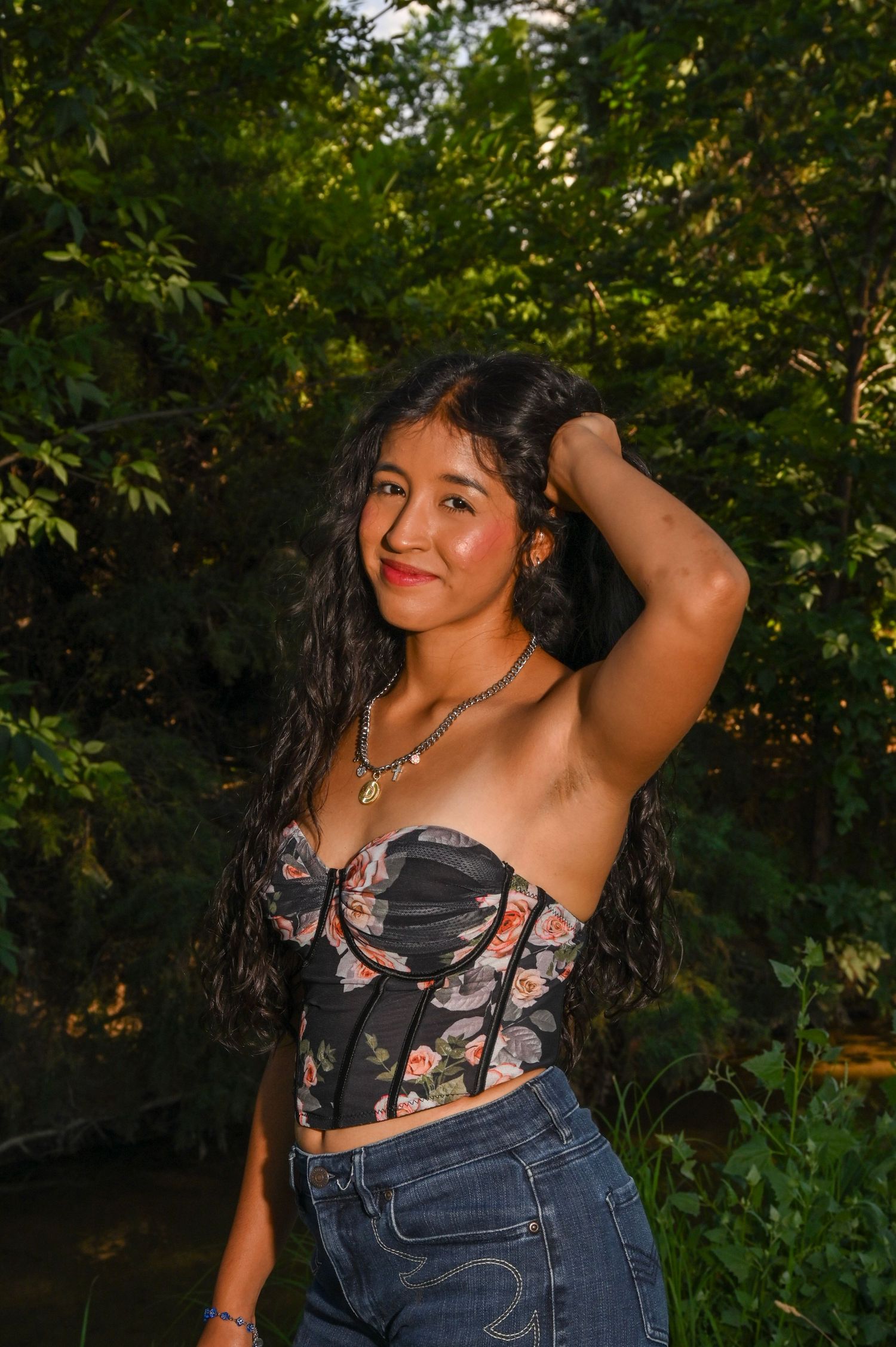 Young woman in floral corset top and jeans posing outdoors among lush green trees at golden hour.