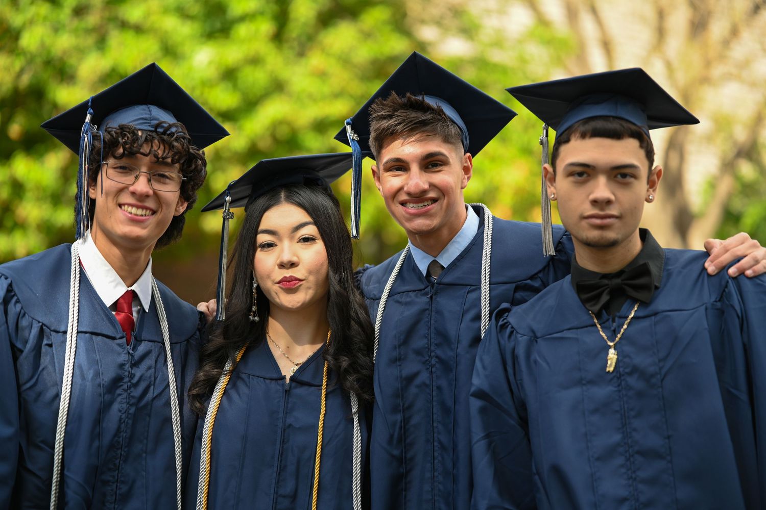 Four smiling graduates in navy blue caps and gowns pose together outdoors at a graduation ceremony.
