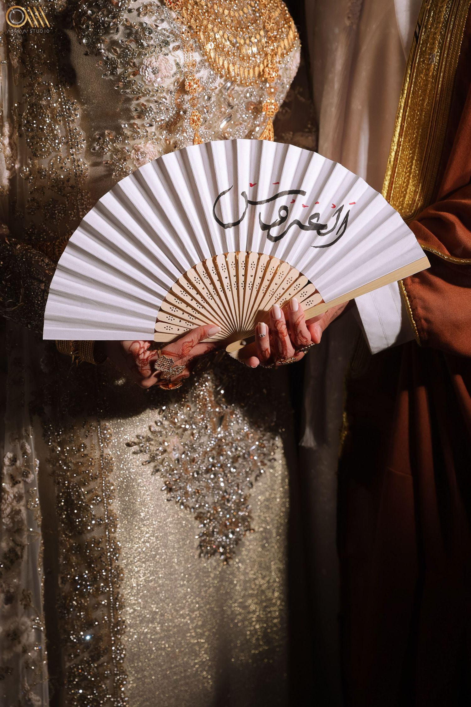 Ornate white folding fan with Arabic calligraphy held against sparkly formal attire.