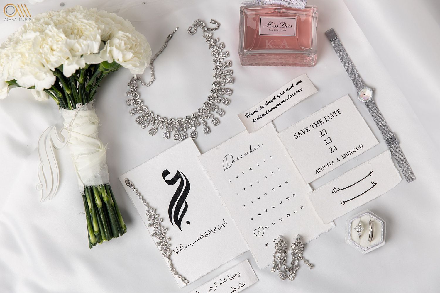 Wedding accessories including perfume, jewelry, flowers and stationery arranged on white surface.