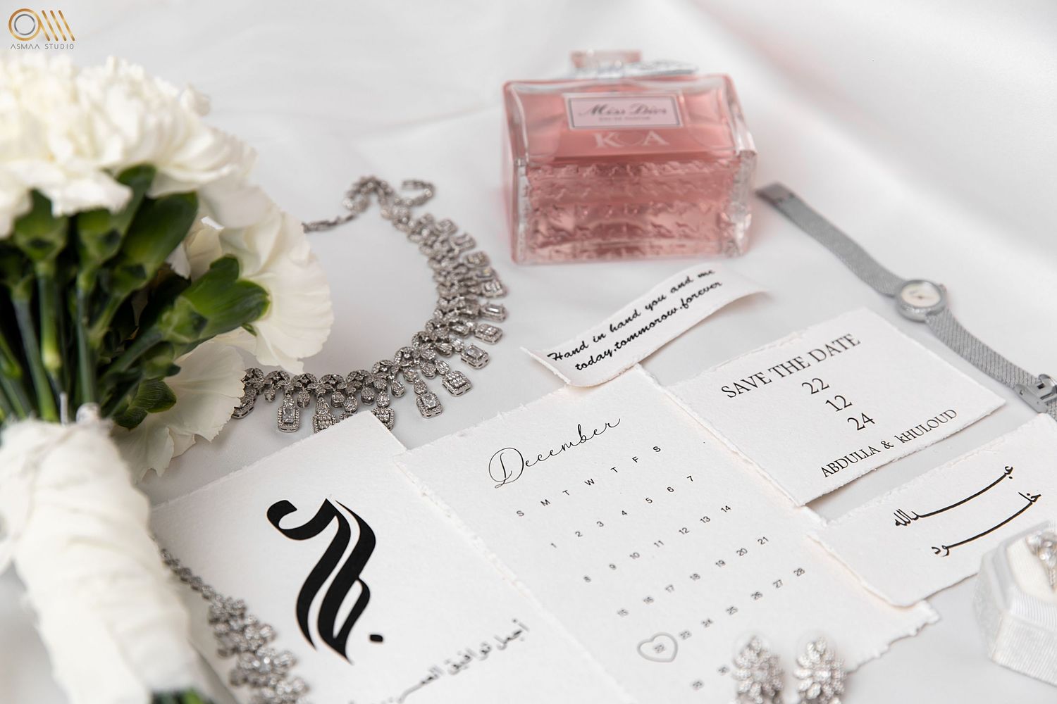 Wedding details flatlay with pink perfume bottle, white flowers, and Arabic stationery.