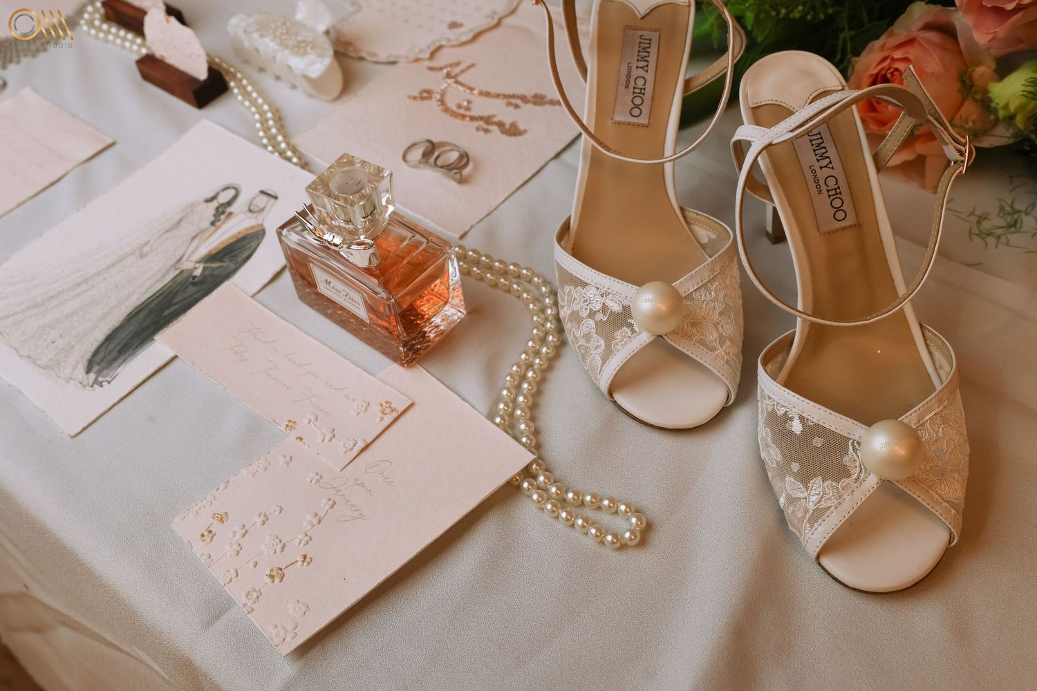 Elegant wedding accessories including white heels, pearl necklace, and invitation cards arranged on a soft surface.