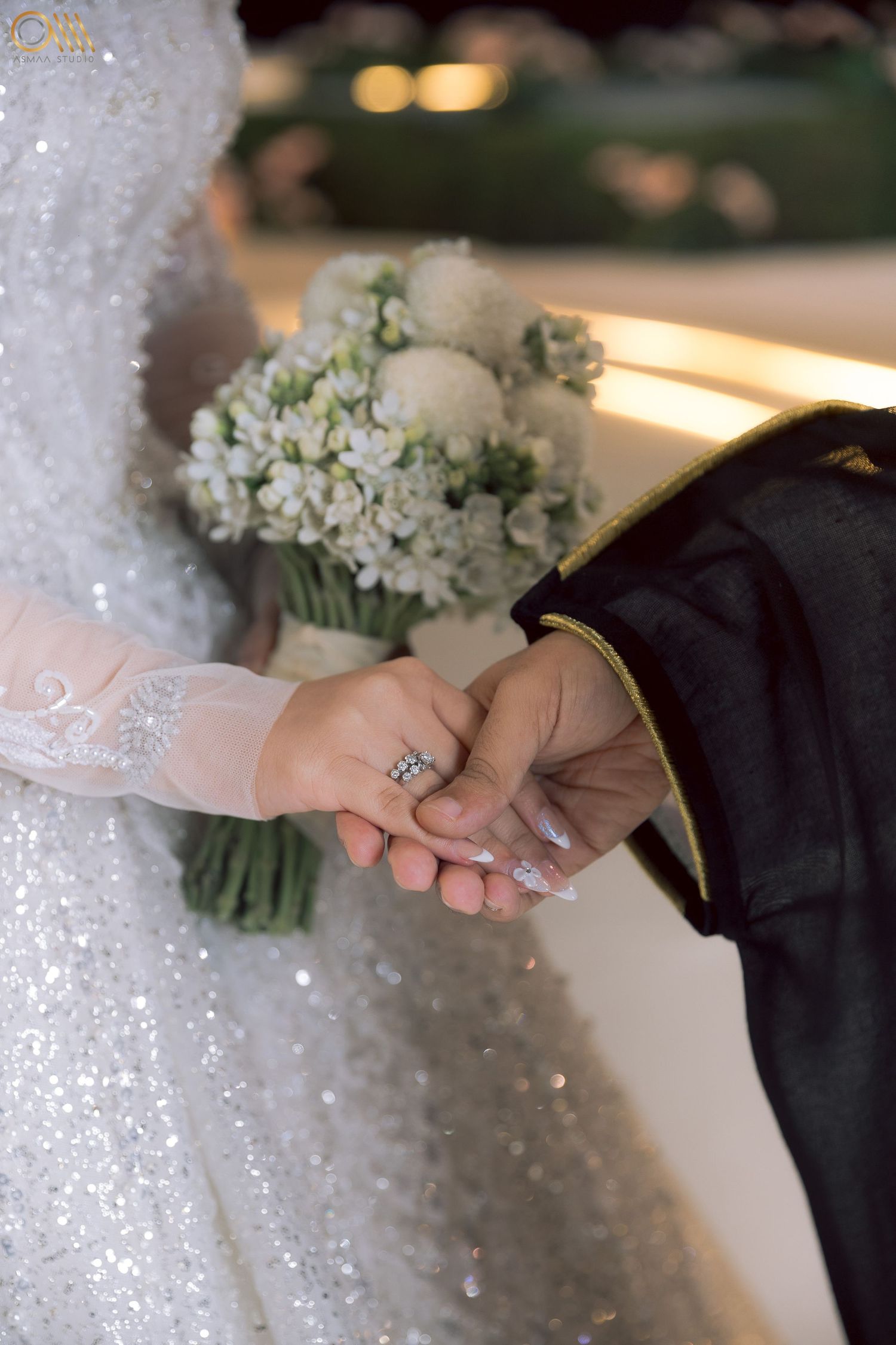 Close-up of hands exchanging rings with sparkly wedding dress and white flowers in frame.