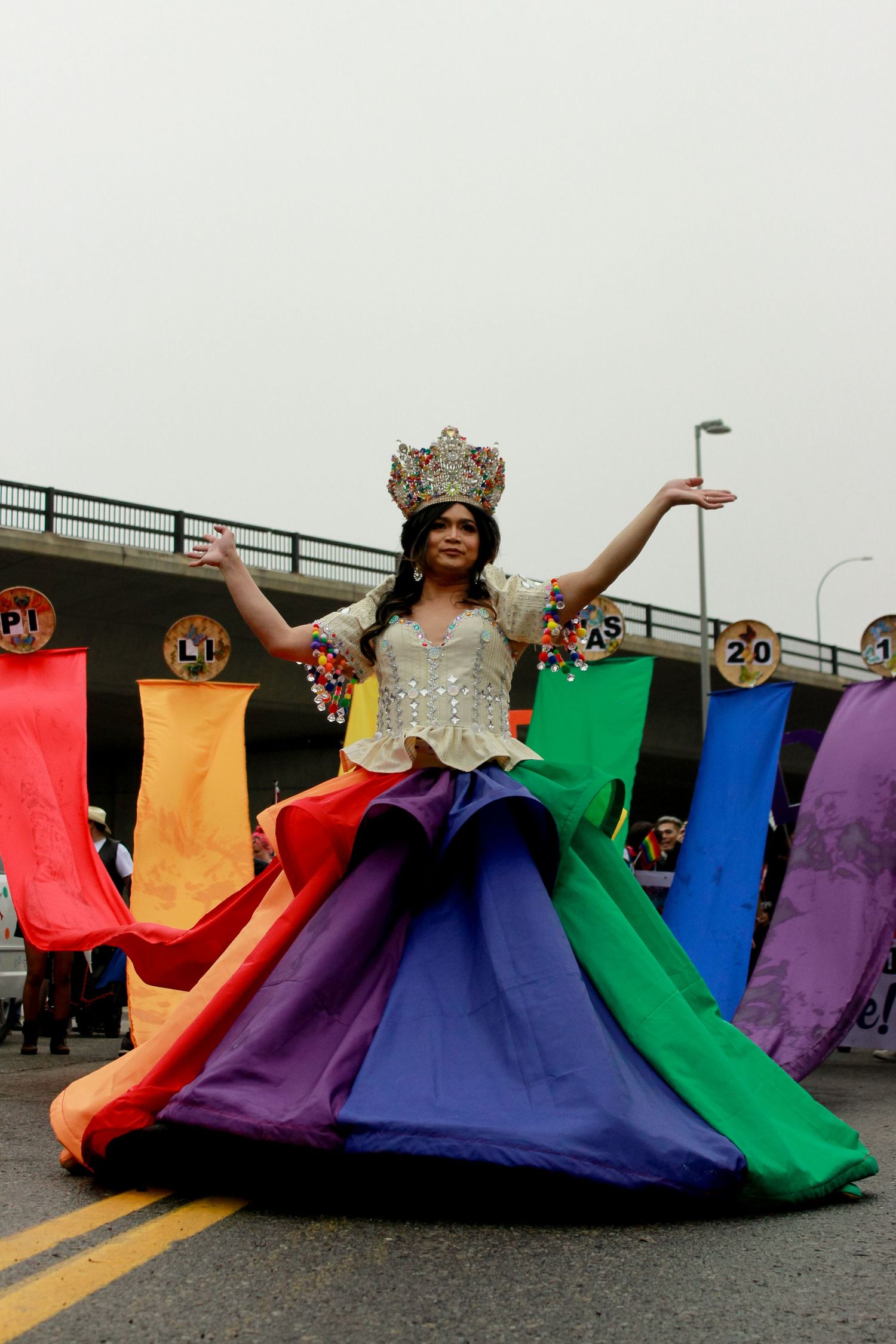 Performer in colorful rainbow dress and crown posing dramatically outdoors.