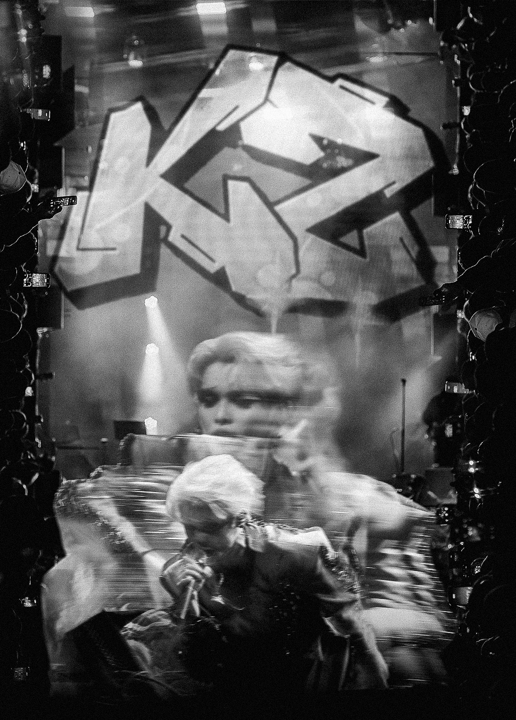 Black and white concert photo with graffiti backdrop showing a blond performer singing into a microphone on stage.