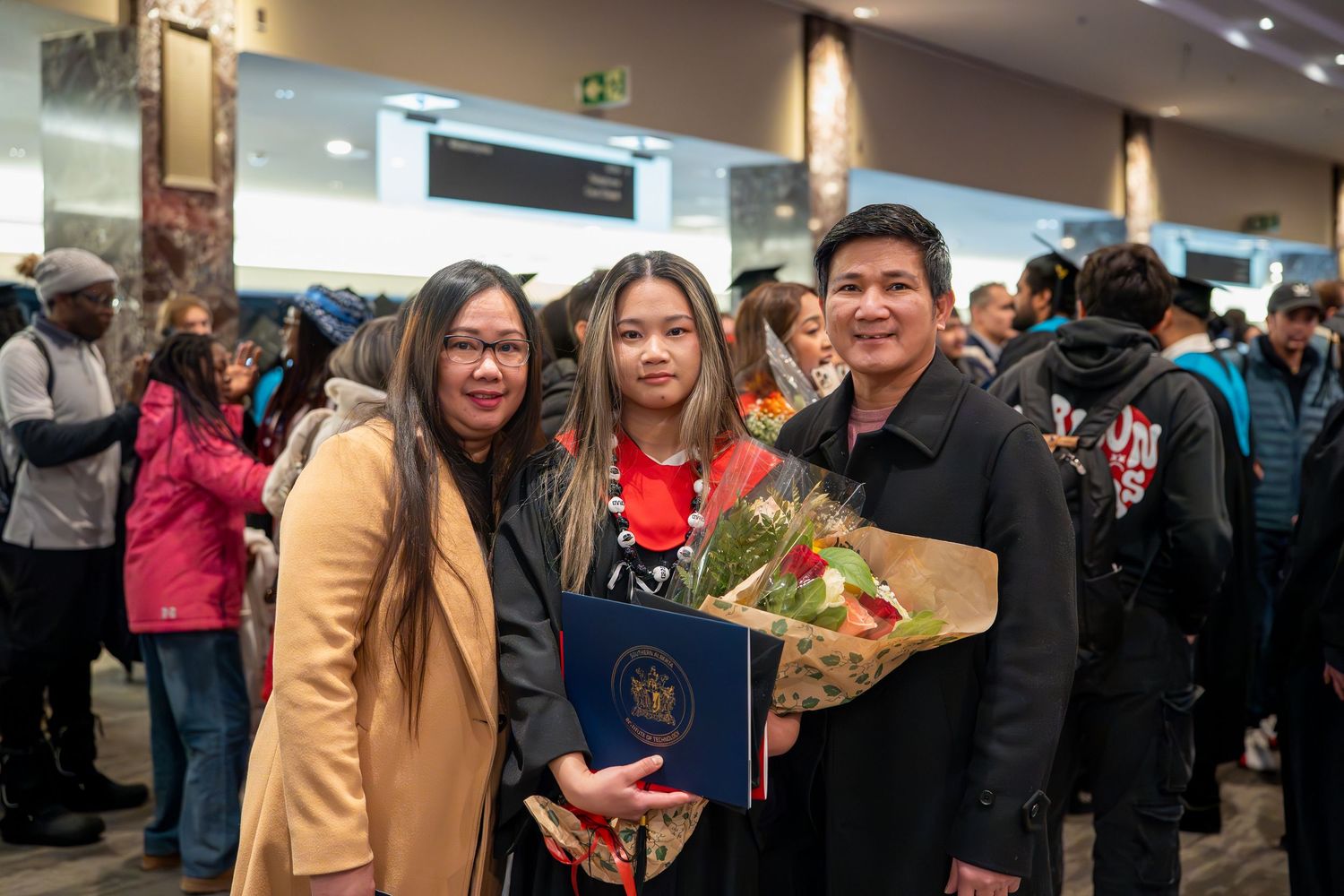 Graduation celebration scene with graduates holding diplomas and flowers in crowded venue.