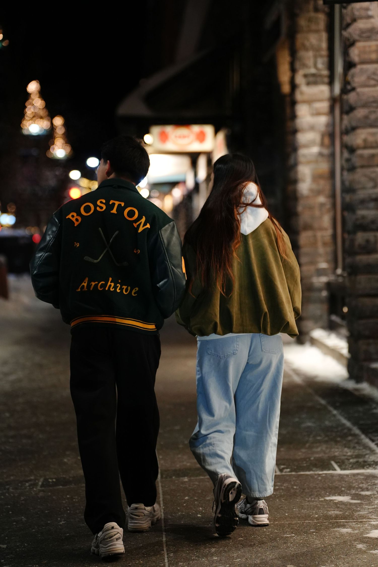 Person wearing Boston Archive jacket walking with companion on nighttime street.