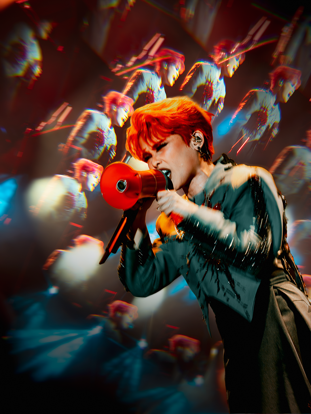 A vibrant performer with bright orange hair sings into a red megaphone on a dramatic, bokeh-lit concert stage.