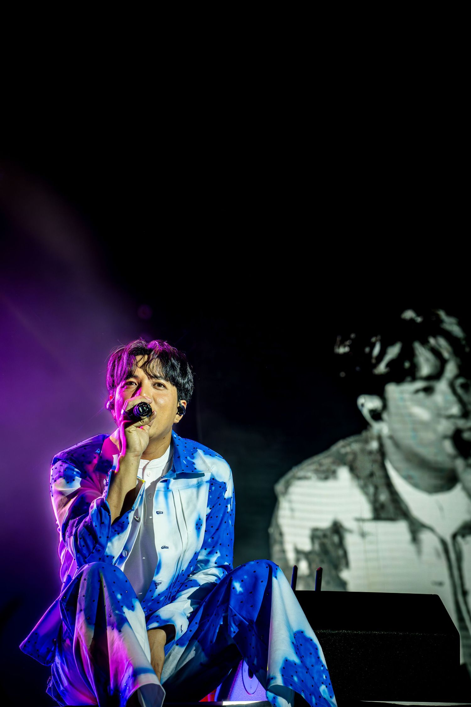 Singer performing on stage under purple lights, wearing a blue tie-dye outfit, with a projected image behind them.