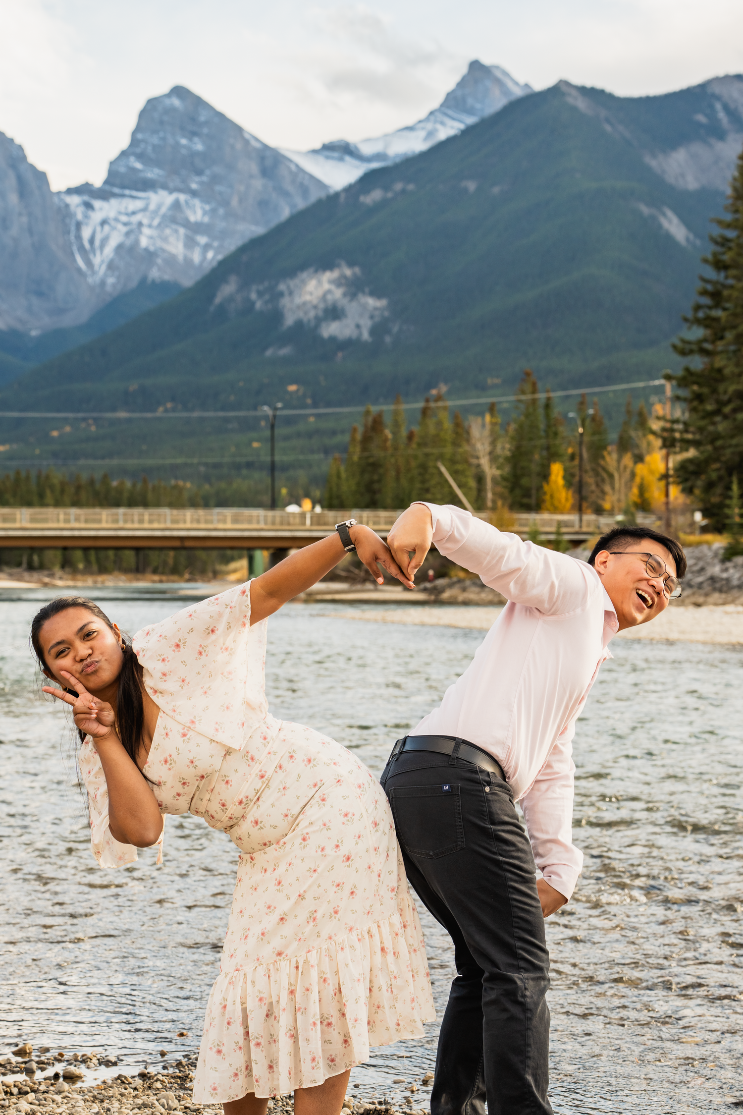 A playful couple poses by a lake with dramatic mountain peaks in the background.