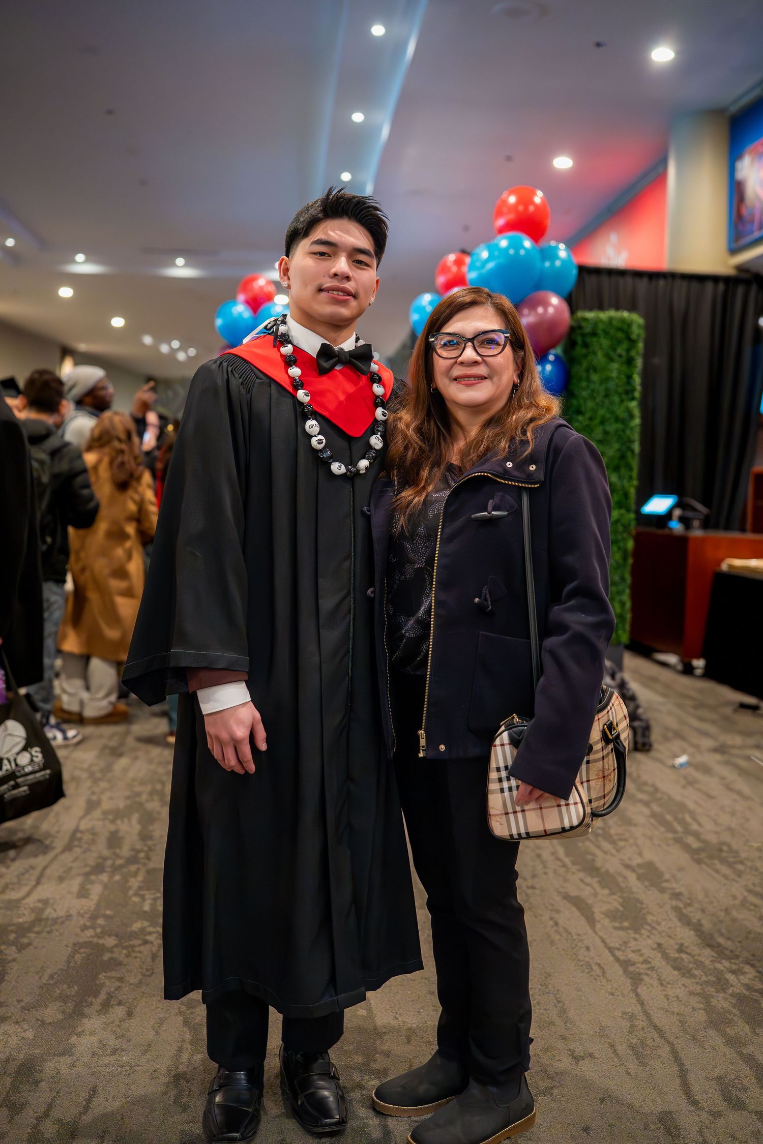 Graduate in academic regalia posing with family member at graduation ceremony with balloon decorations.