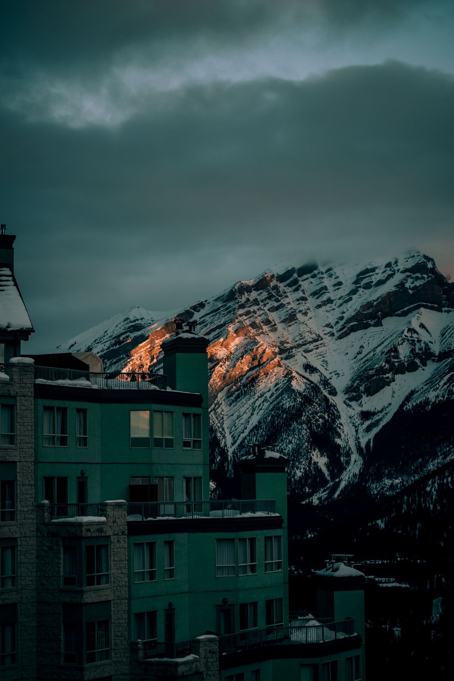 Snow-capped mountain peaks at dusk with modern buildings in the foreground.