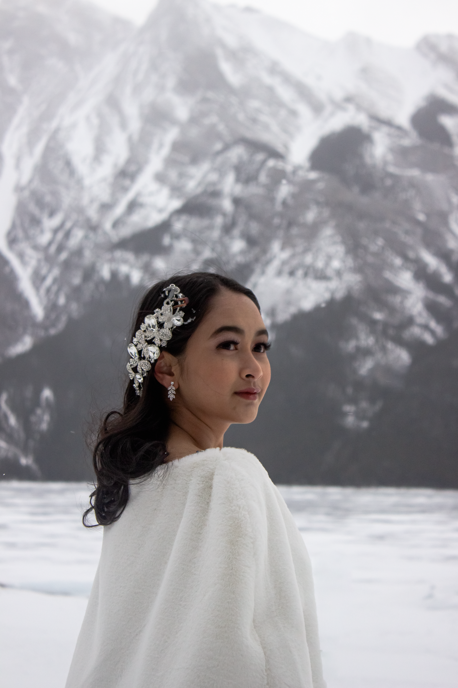 Winter portrait against snowy mountain backdrop featuring white fur wrap and crystal hair accessories.