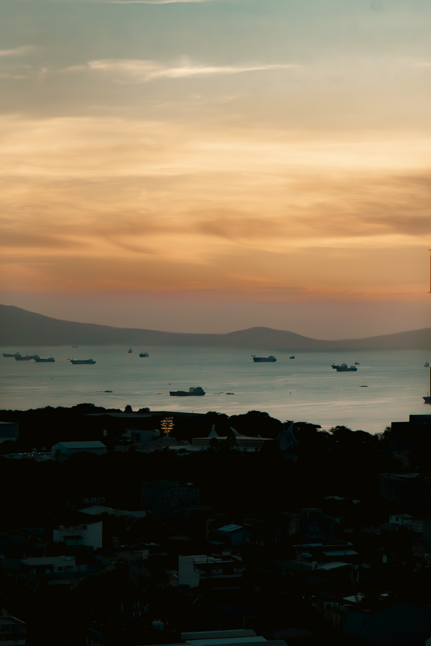Sunset over coastal city silhouette with ships visible on horizon.