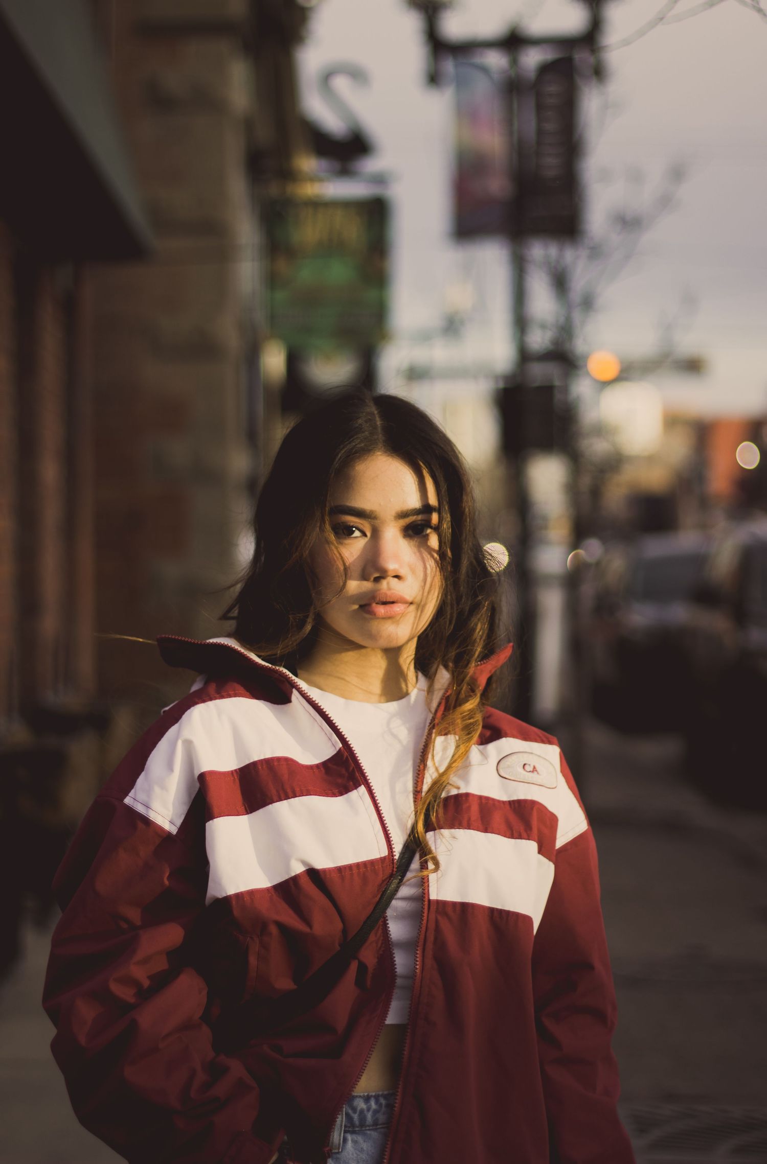 Person wearing a burgundy and white jacket poses on a city street at dusk with soft lighting.