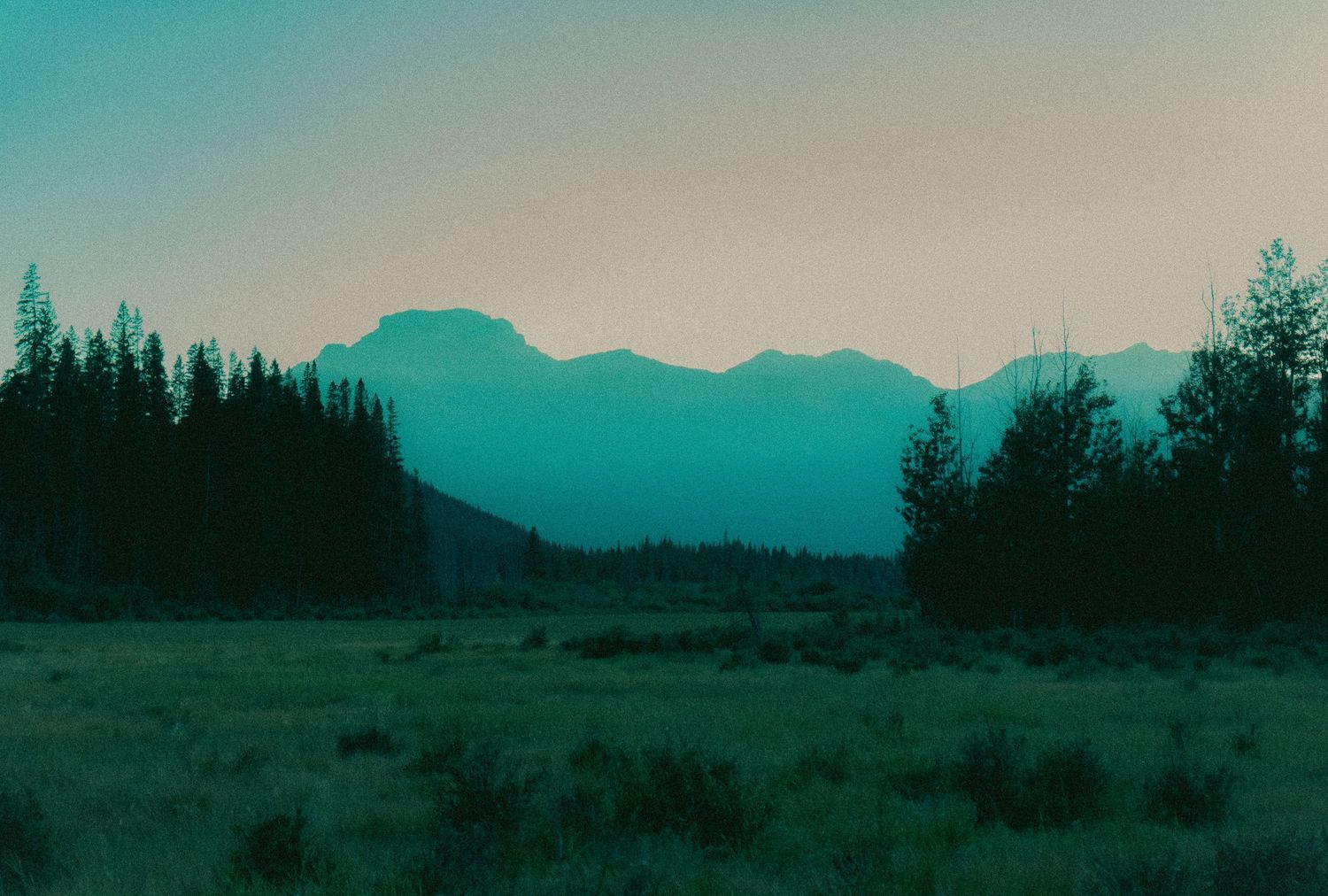 Panoramic mountain landscape at dusk with silhouetted treeline against turquoise sky.