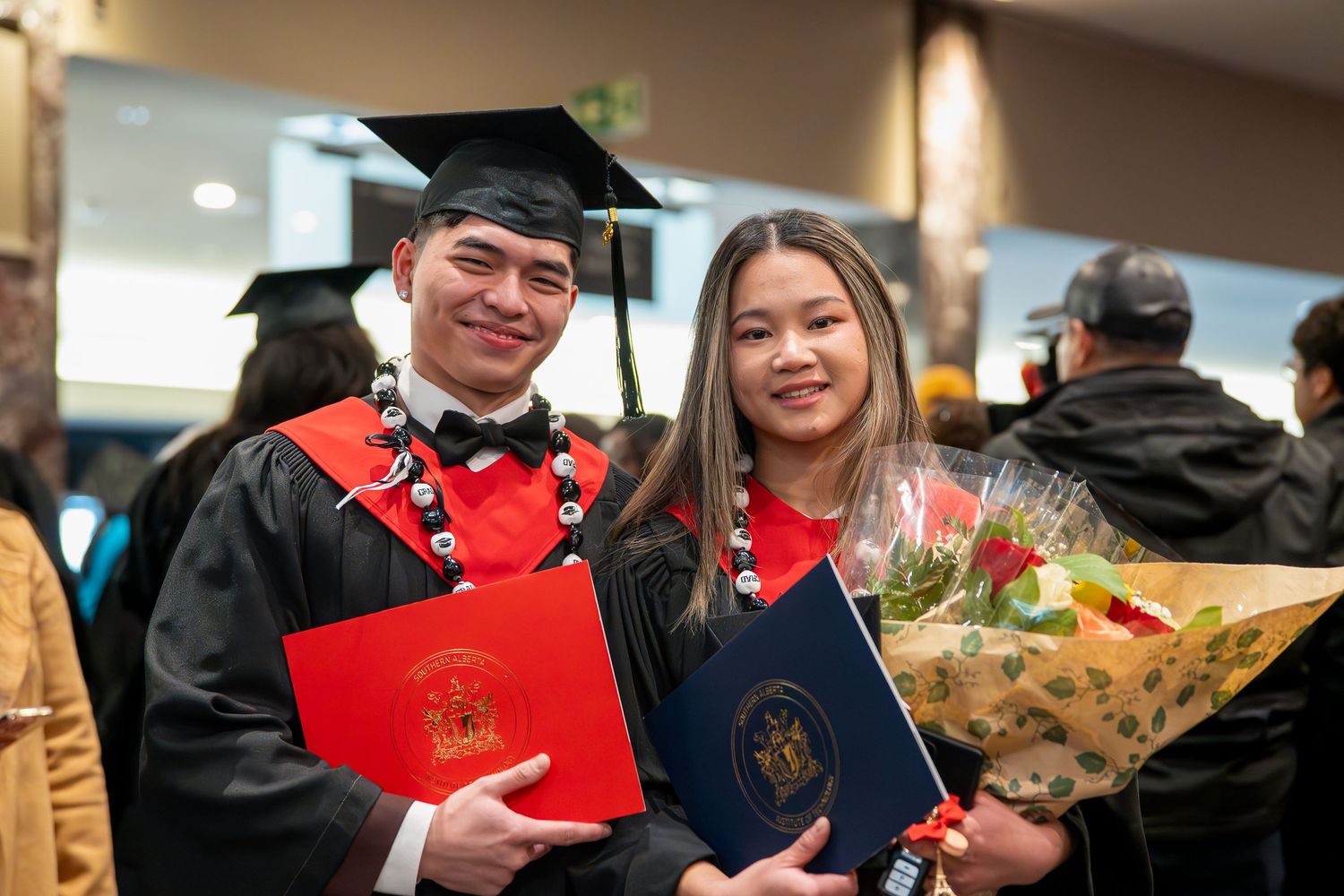Graduates in academic regalia holding diplomas and flowers at commencement ceremony.