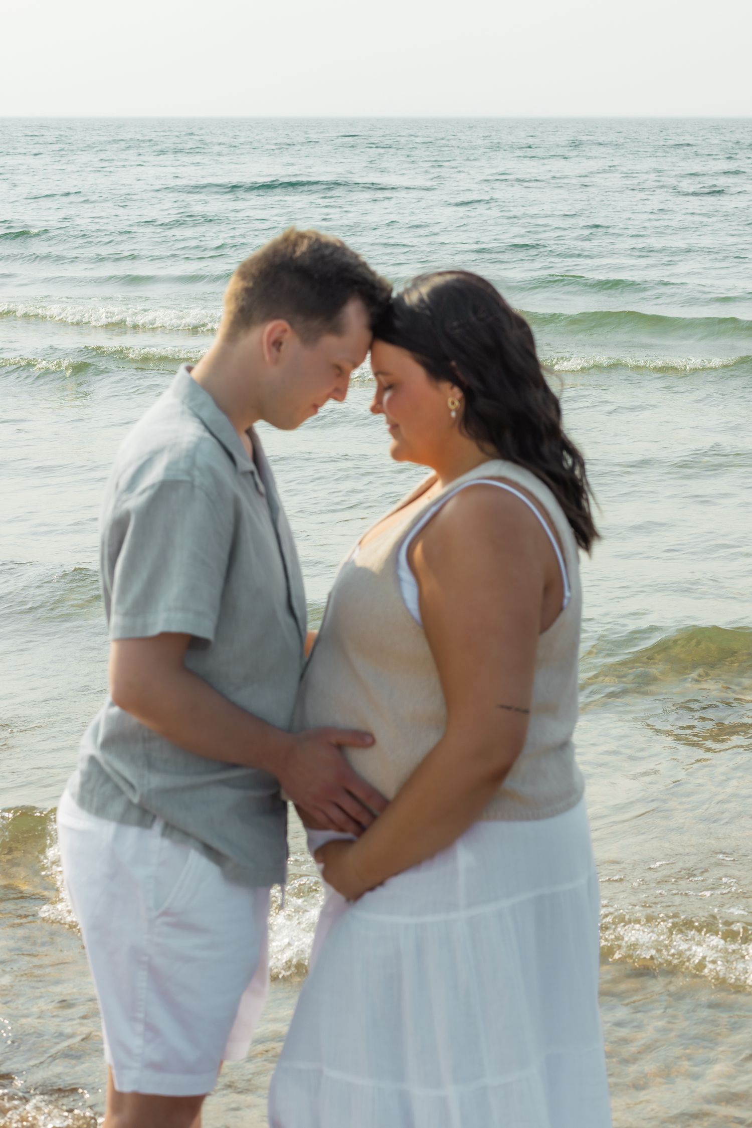 Pregnant couple sharing tender moment on beach, foreheads touching while holding baby bump together.