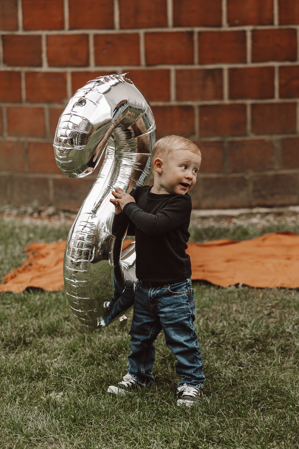 Toddler holding large silver number 2 balloon against brick wall background.