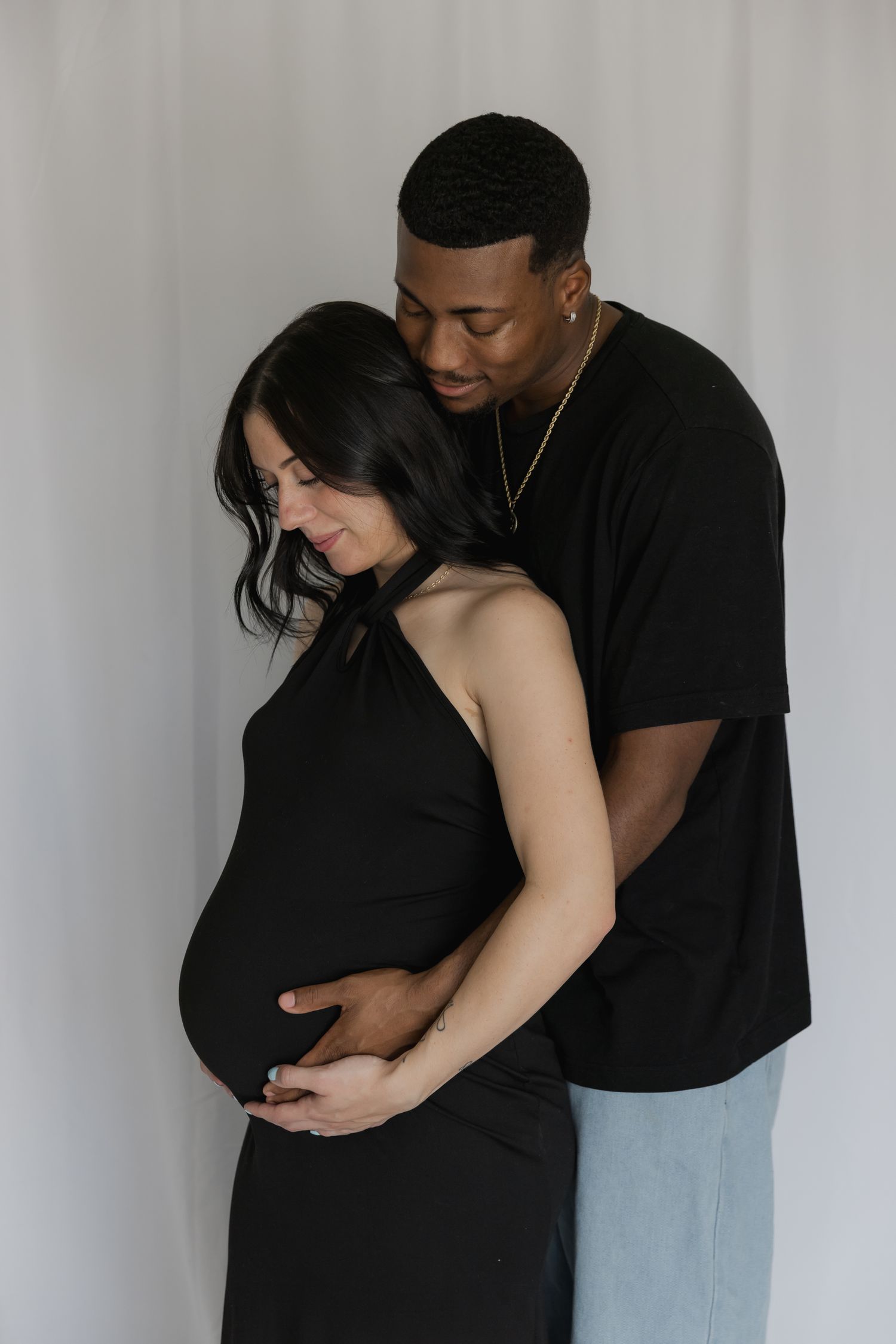 A couple shares a tender maternity photo, the pregnant woman in black dress embraced from behind by her partner against a white backdrop.