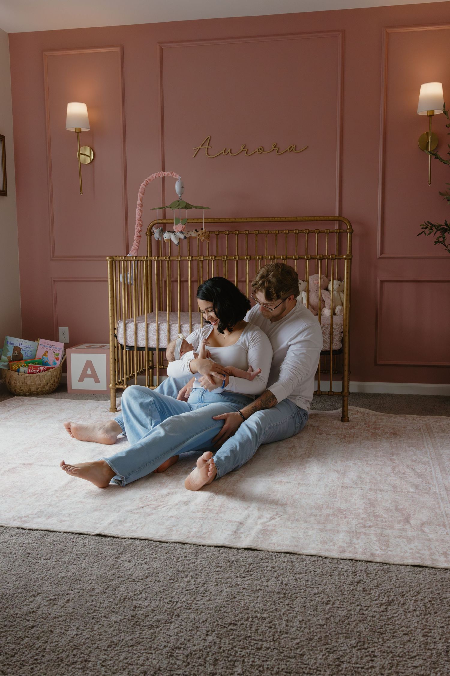 Parents sitting on nursery floor with newborn baby in front of gold crib in pink room named Aurora.