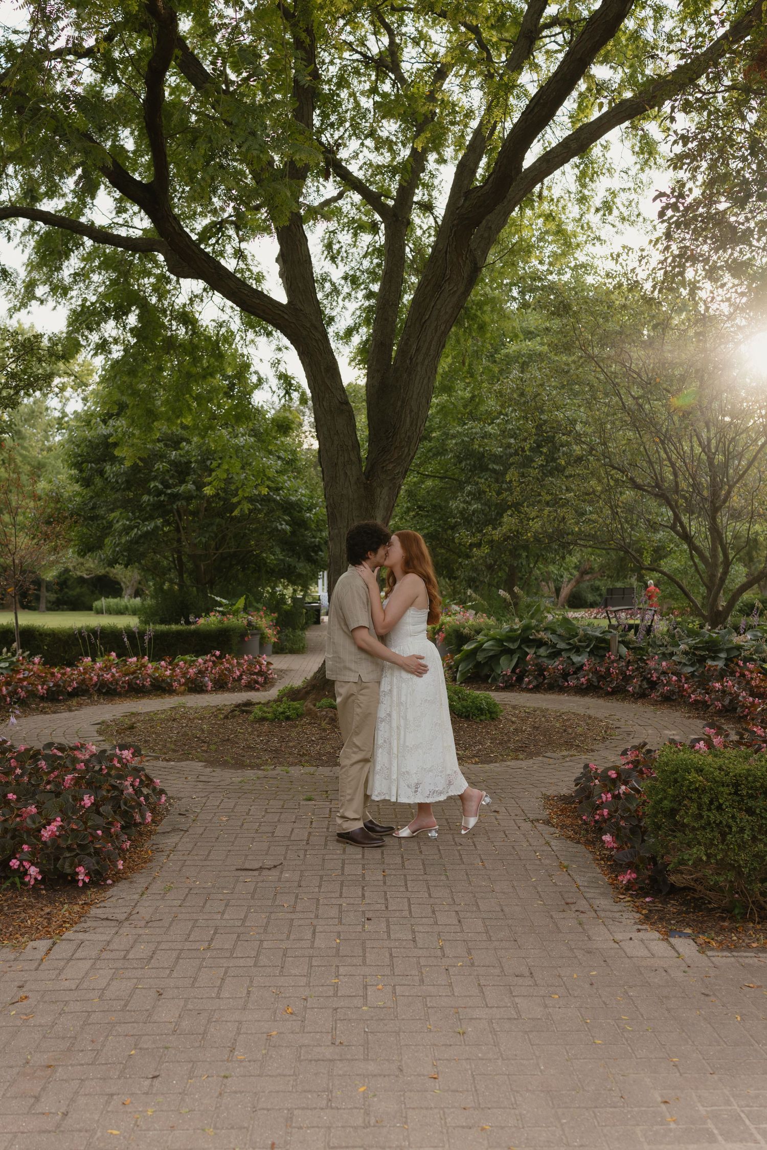 Couple sharing a romantic kiss in a sunlit garden park, surrounded by blooming flowers and a large oak tree.