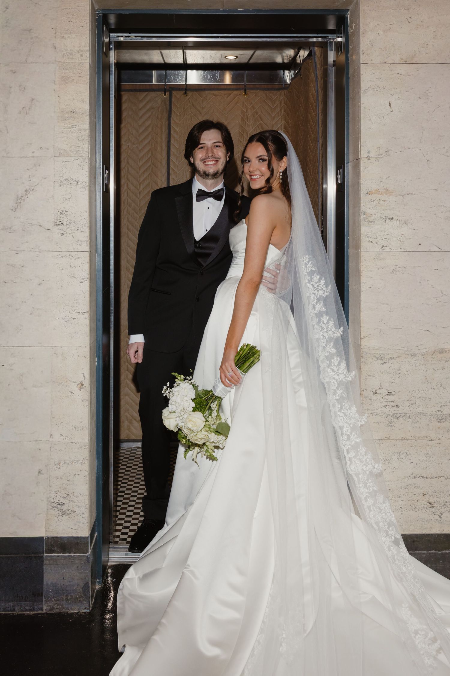 Bride in strapless ballgown with veil and groom in black tuxedo posing in elevator doorway on wedding day.