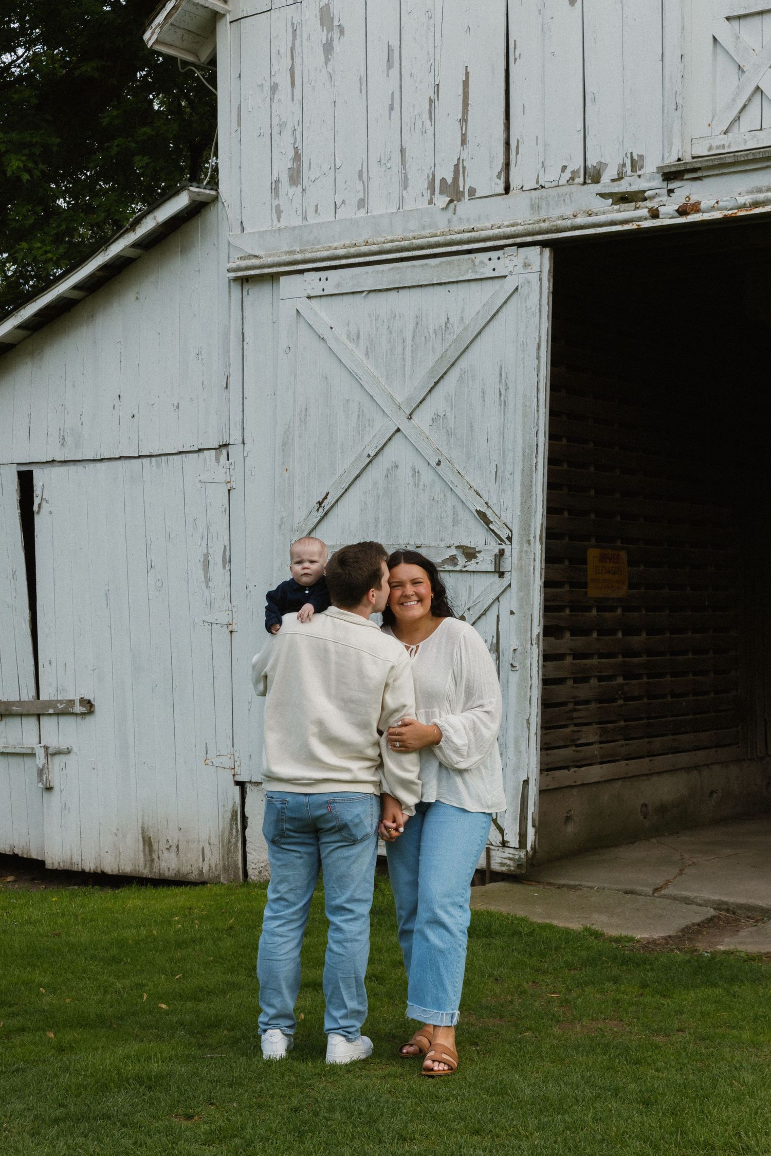 A young family poses in front of a rustic white barn, parents in cream sweaters and jeans holding their baby outdoors.