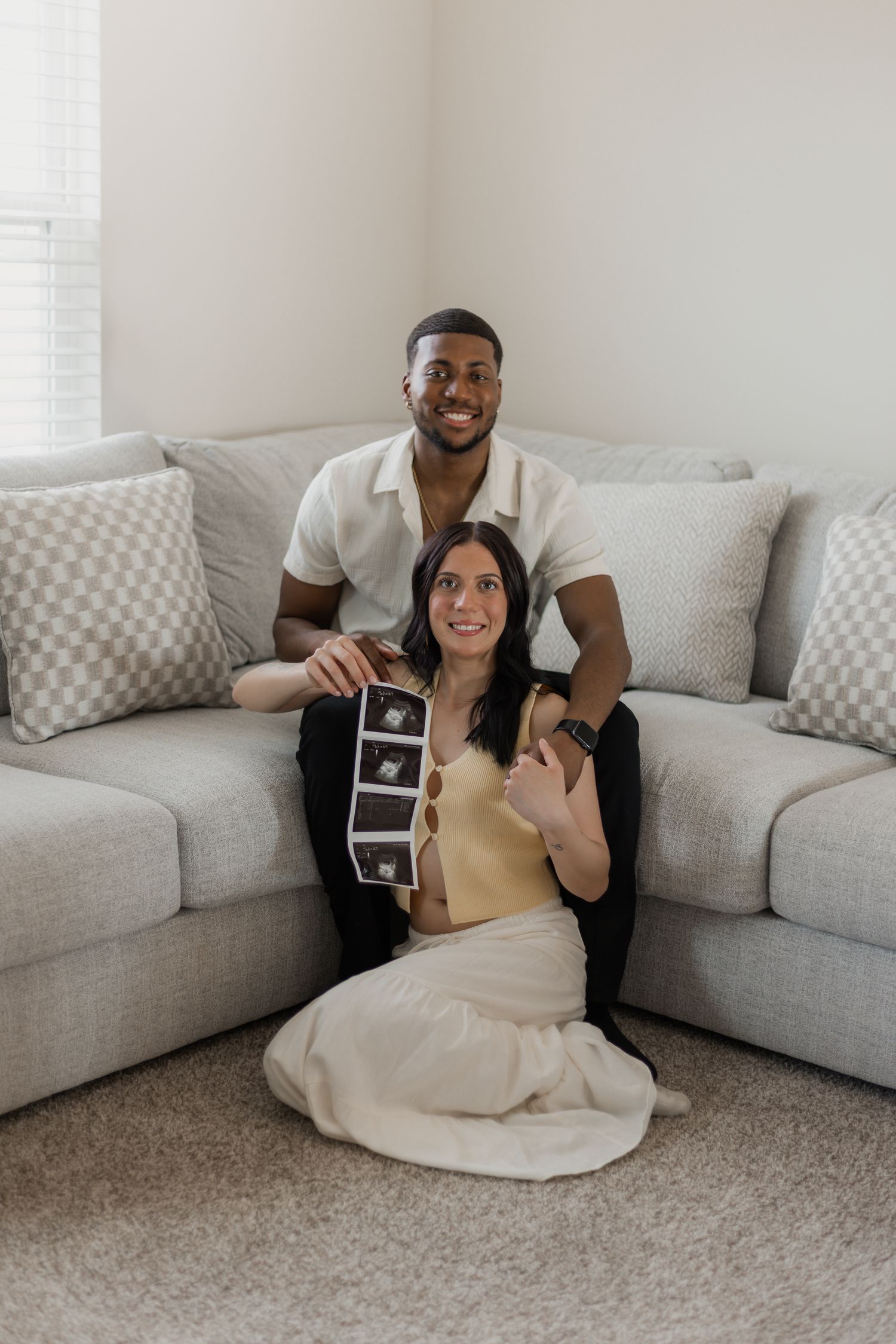 A couple sits on a grey couch holding ultrasound photos while sharing a joyful moment.