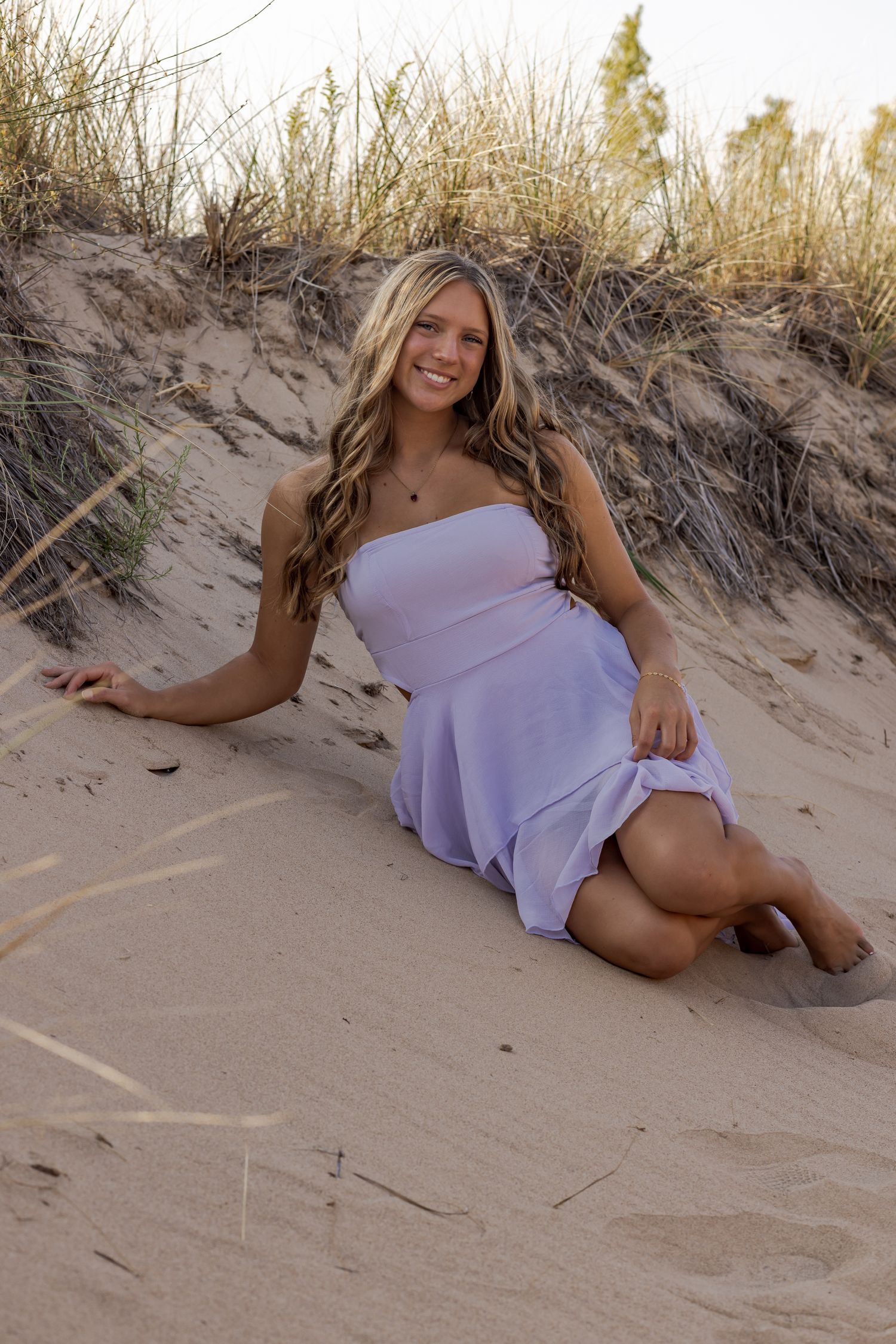Woman in lavender dress lounging on sandy beach dunes at sunset.