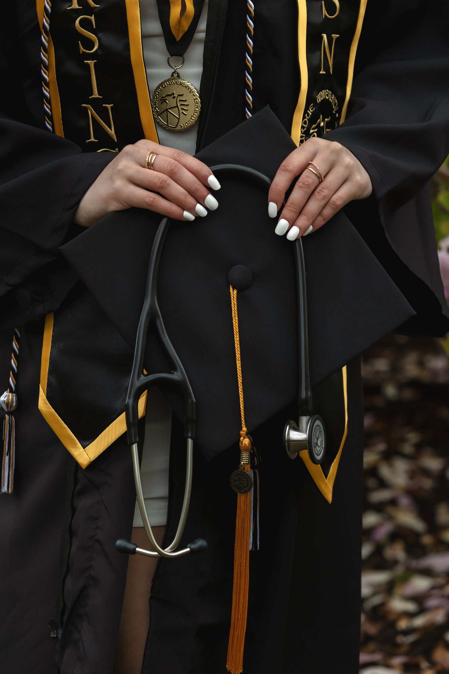 Nursing graduate in black and gold gown holding graduation cap with stethoscope and honors medals.