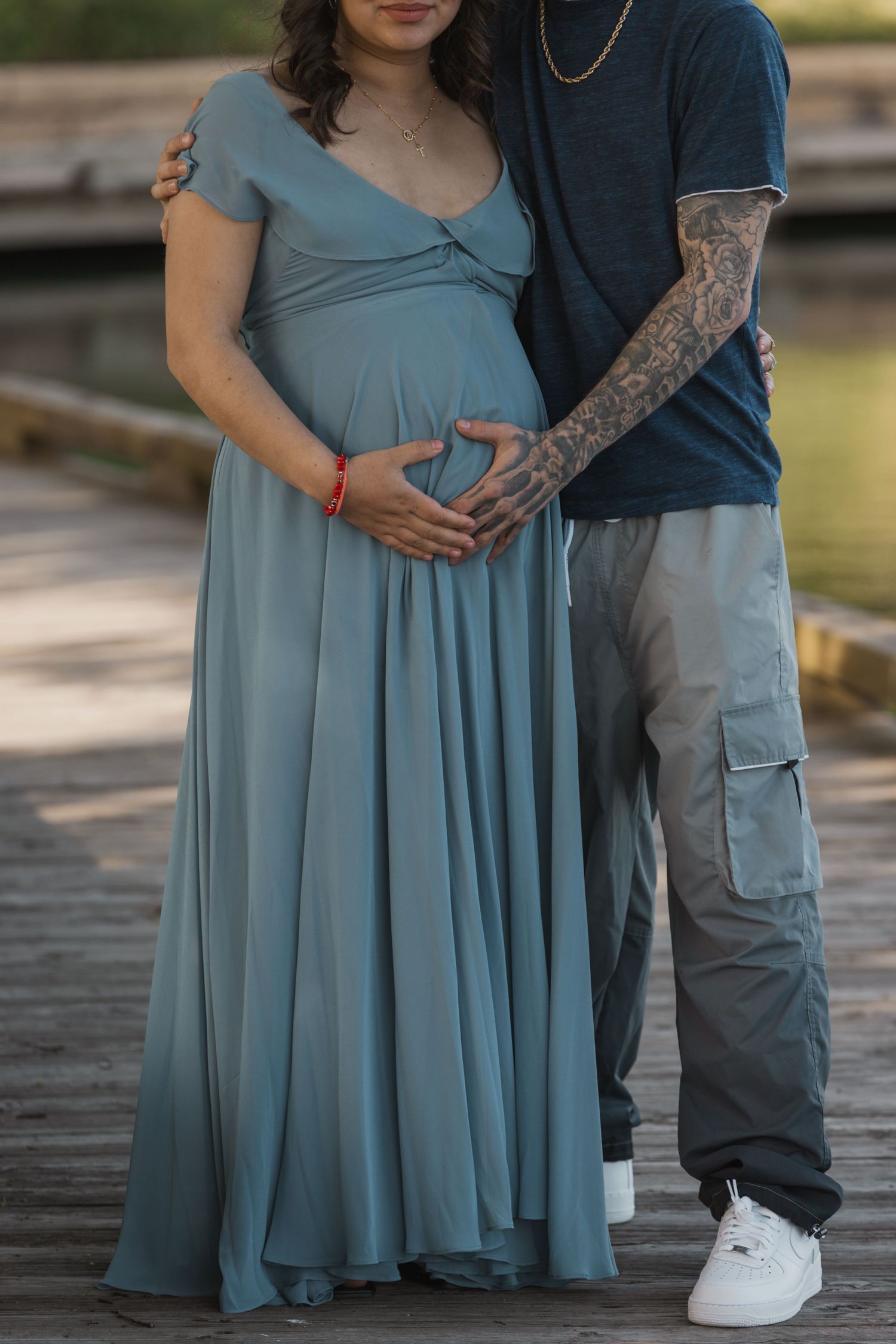 Pregnant couple in teal maternity gown and tattooed partner cradling baby bump on wooden dock.