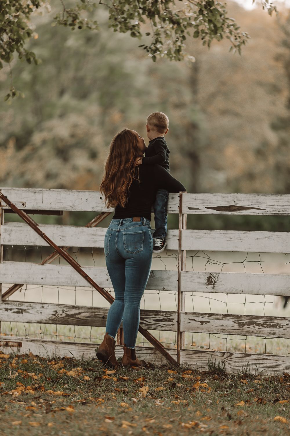 A mother holds her toddler while standing by a rustic white wooden fence in an autumn countryside setting.