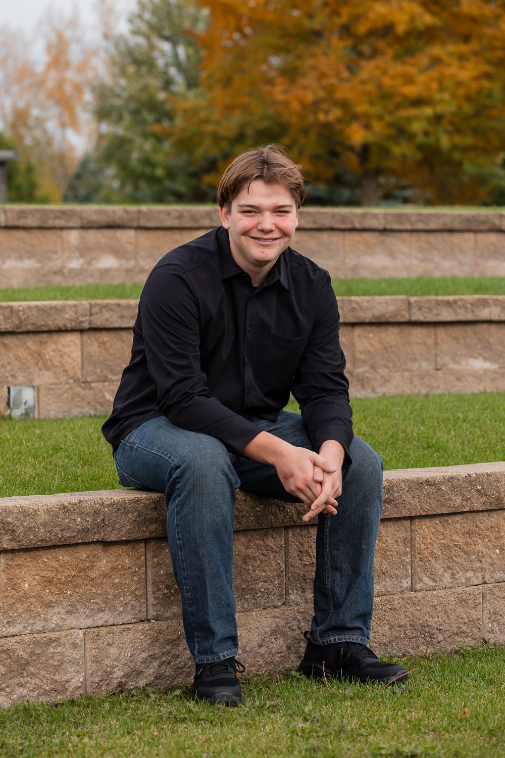 Young man in black shirt and jeans sitting on stone wall outdoors with autumn foliage in background.
