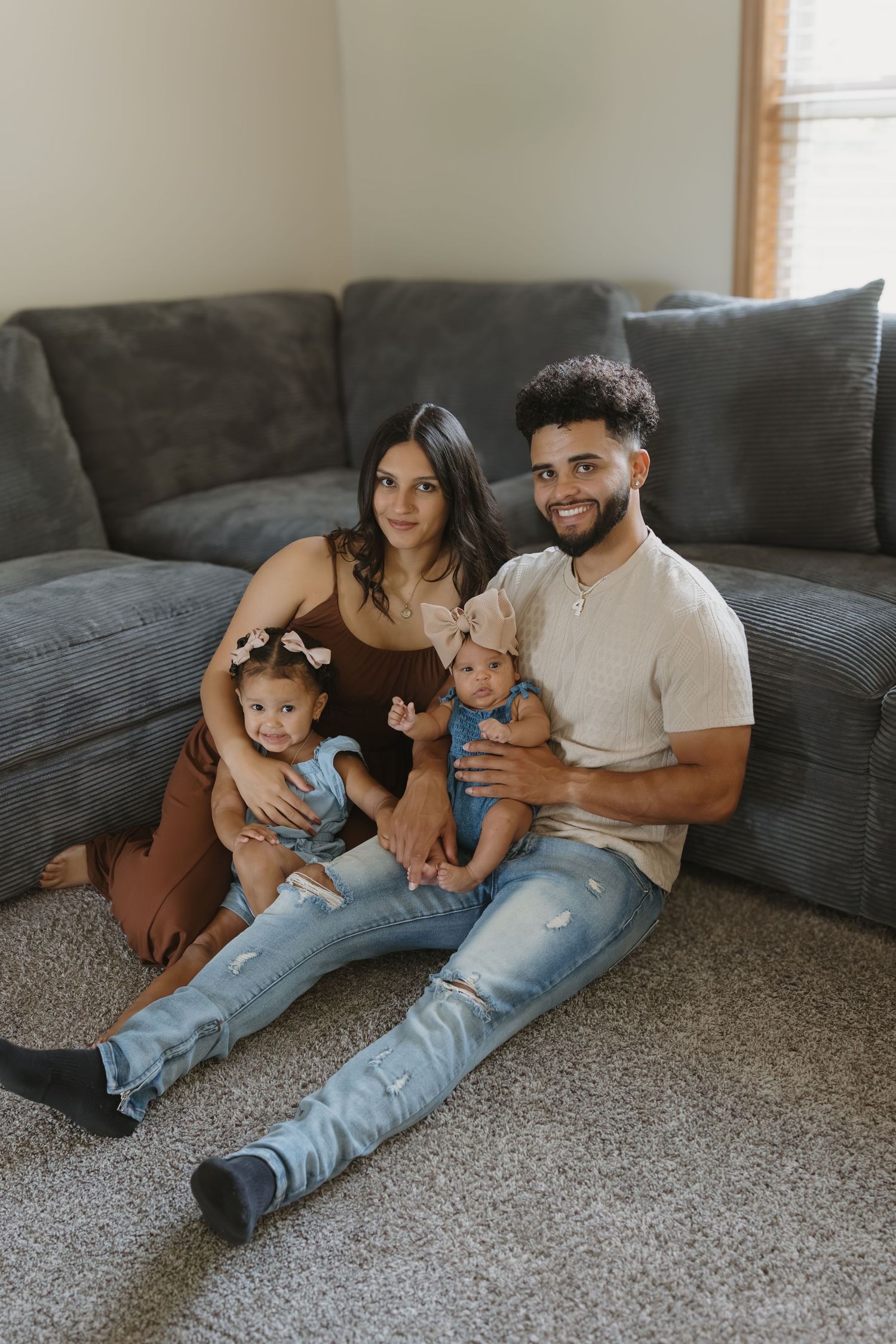 A young family of four sits on the floor against a gray couch, parents smiling while holding their two young children.