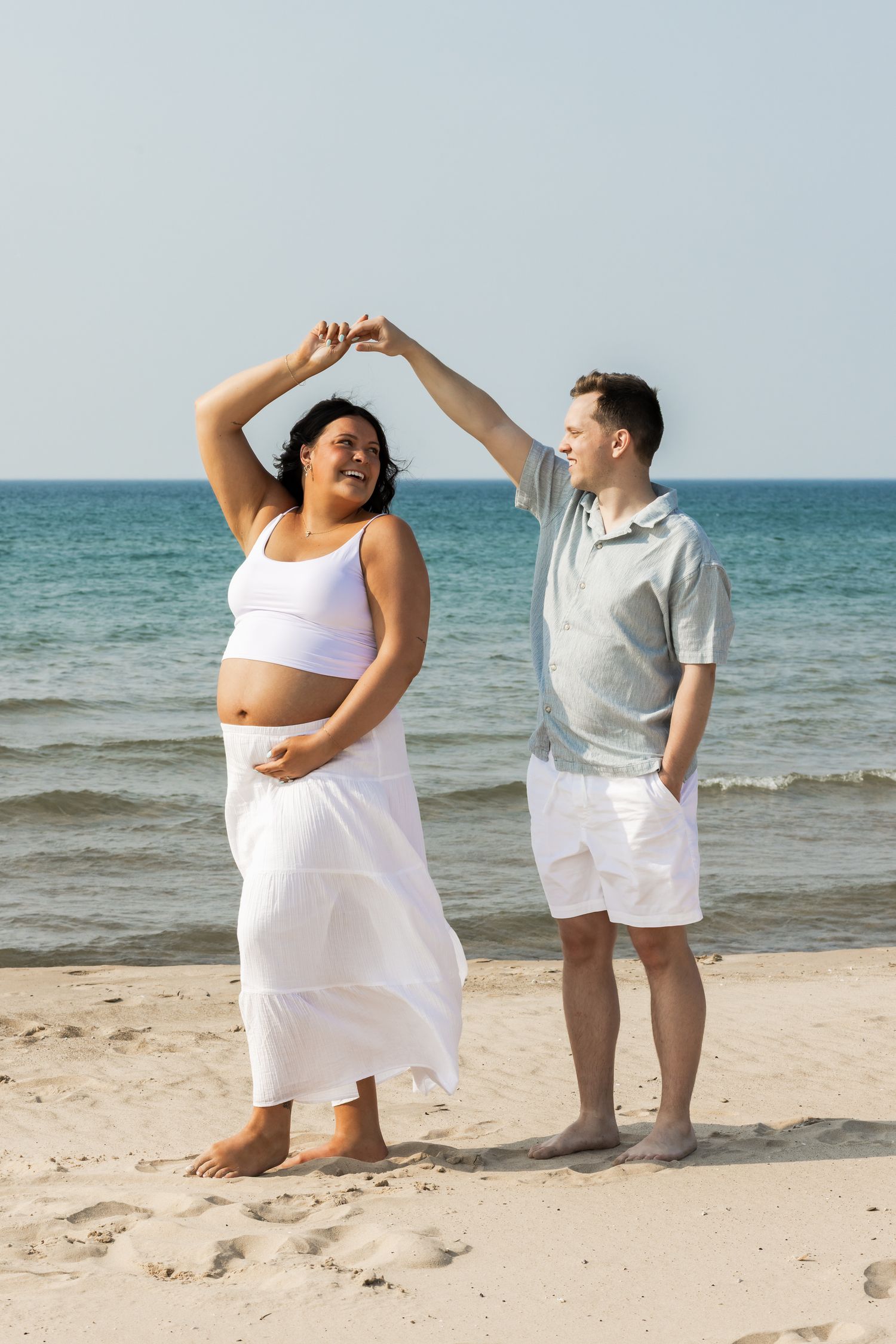 Pregnant woman in white dress dancing with partner on sunny beach, both smiling and holding hands.