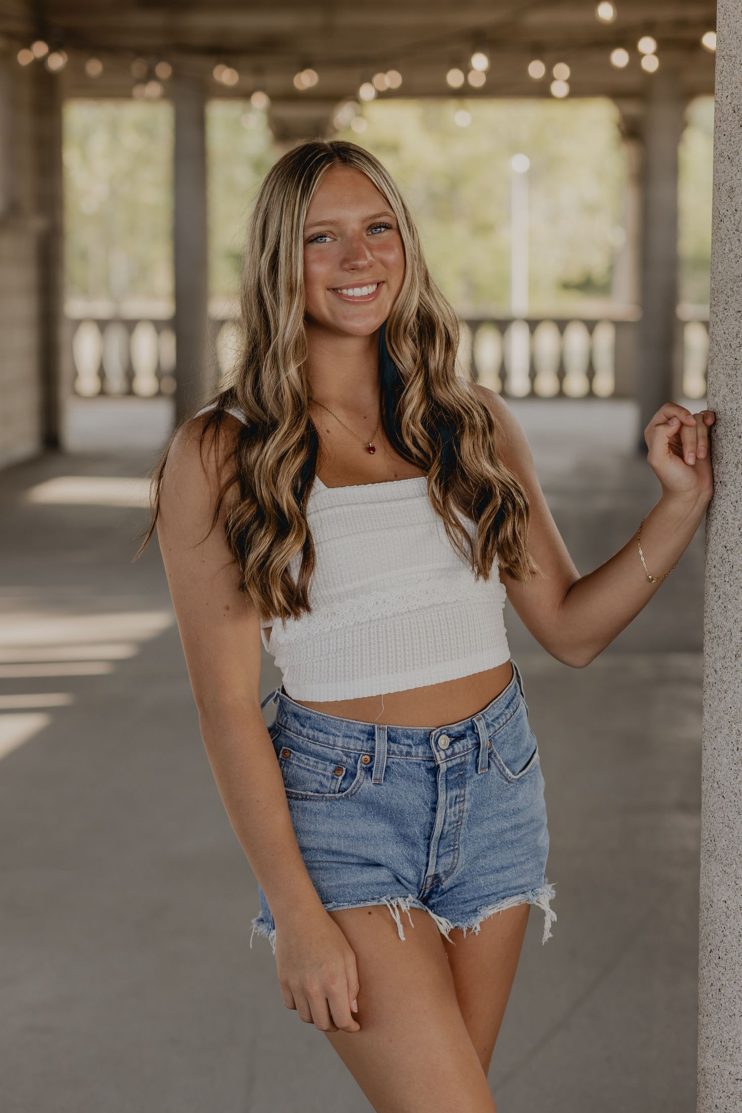 Woman in white crop top and denim shorts poses in a covered outdoor walkway.