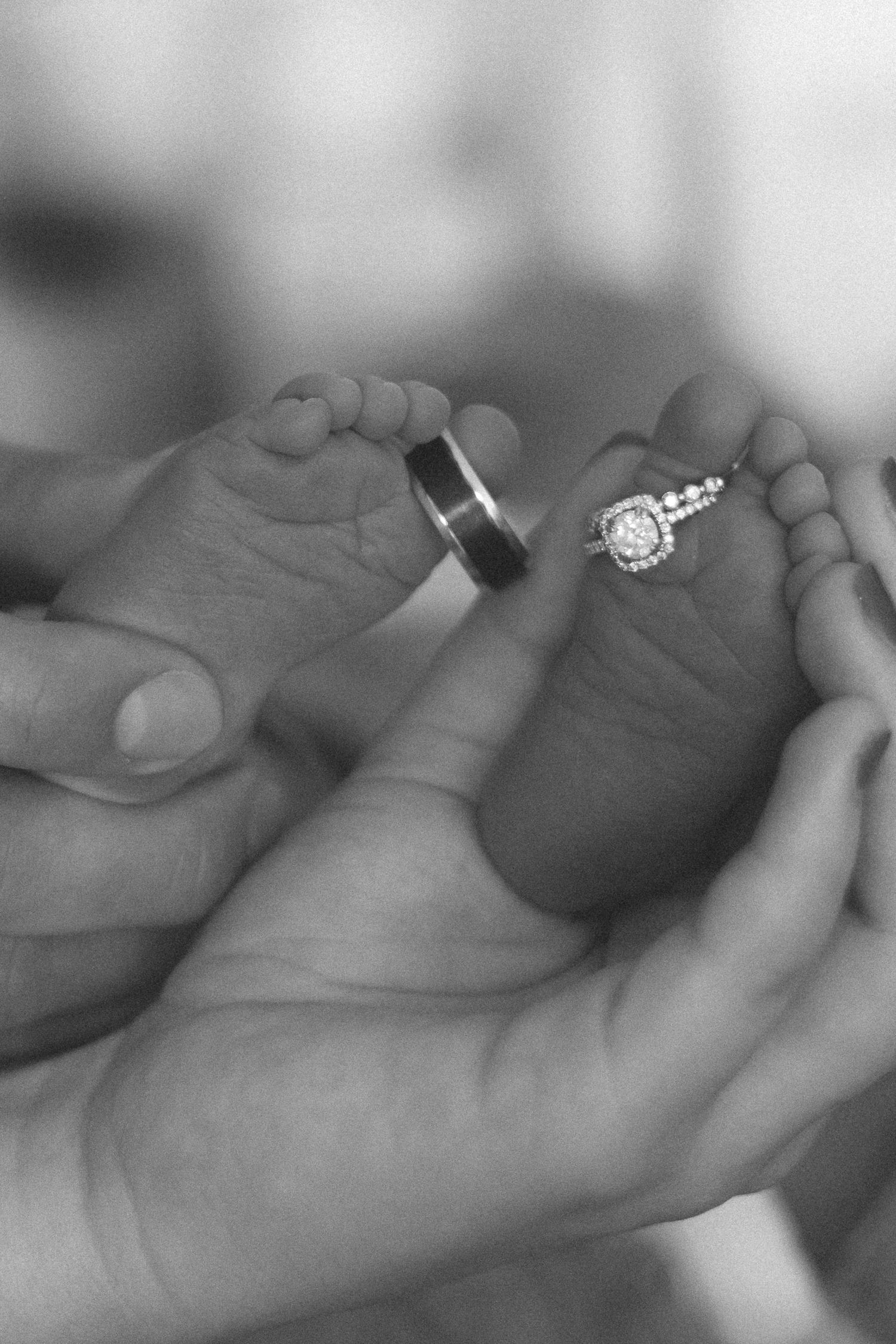 Black and white photo of adult hands cradling newborn baby feet with wedding rings placed on tiny toes.