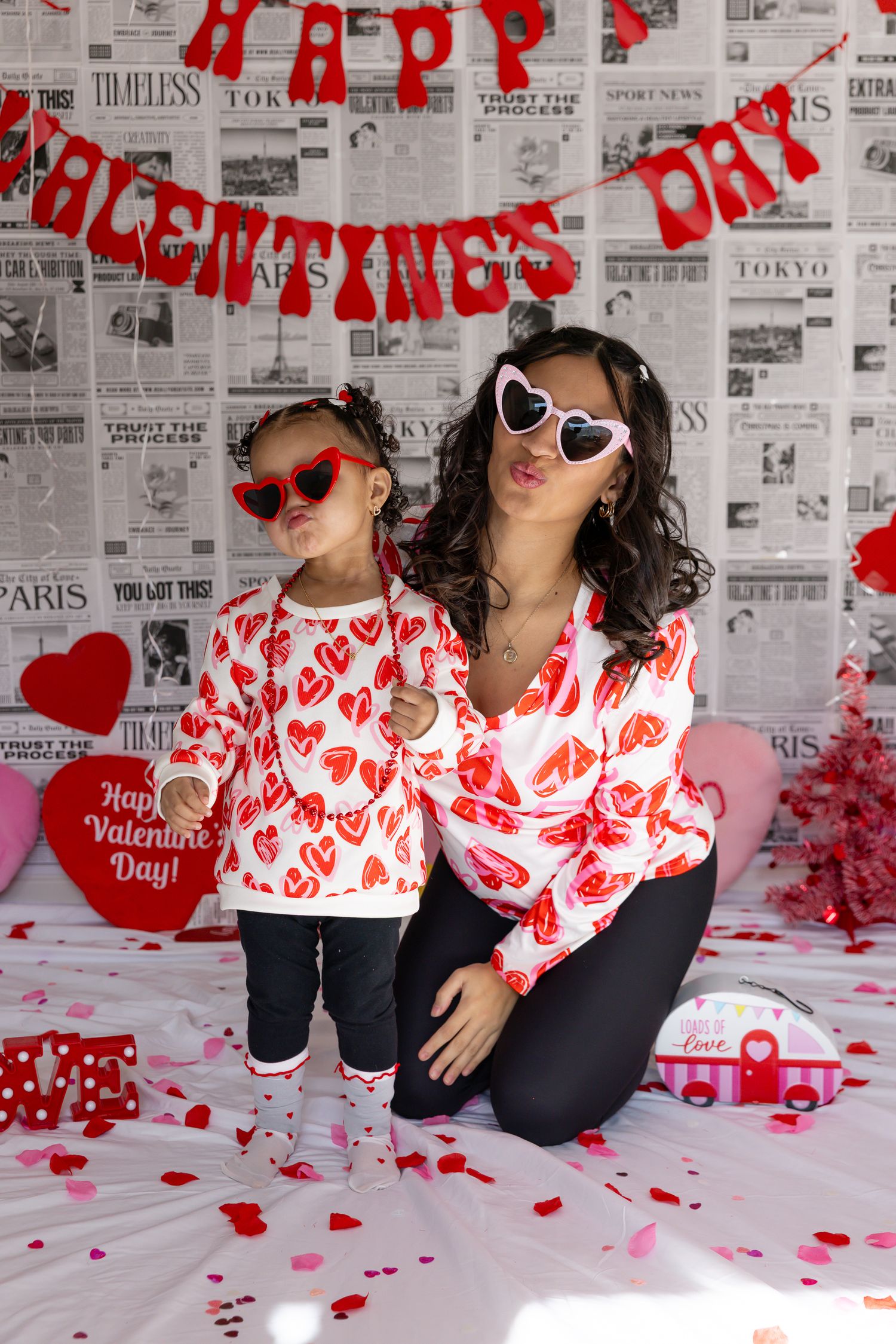Mother and daughter in matching heart-print Valentine's Day outfits and heart sunglasses posing together.