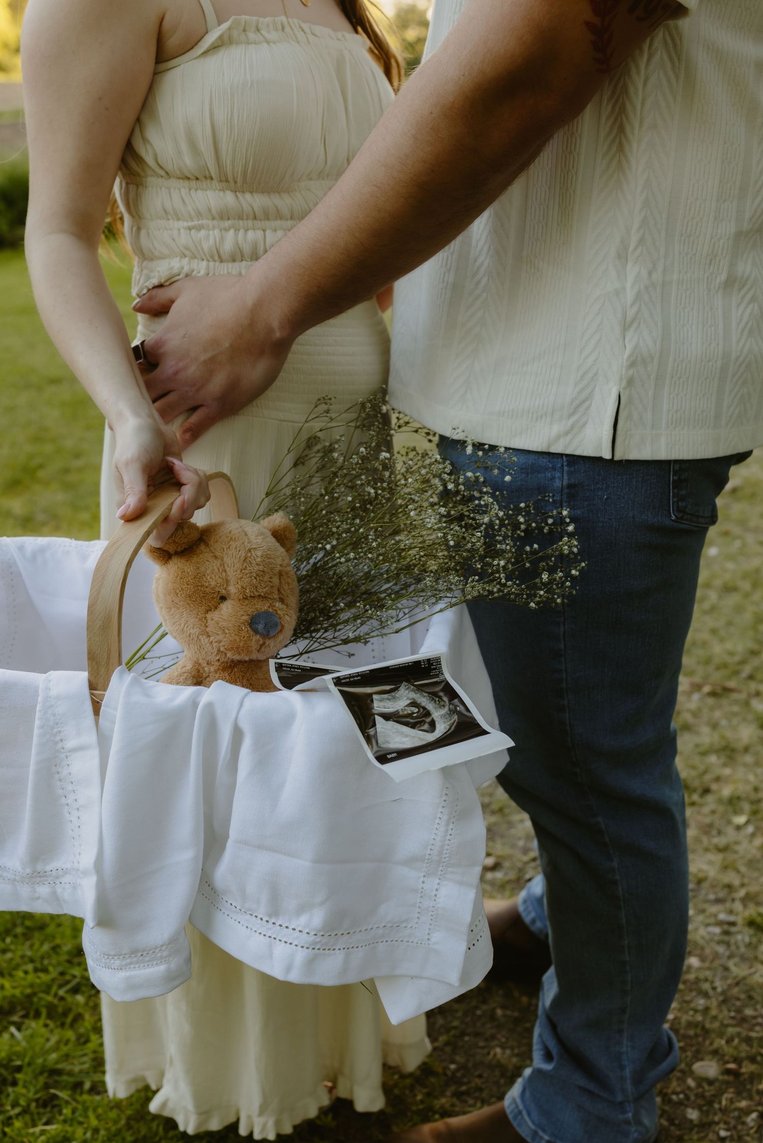 Pregnant couple holding hands outdoors with a basket containing a teddy bear, ultrasound photos, and white blanket.