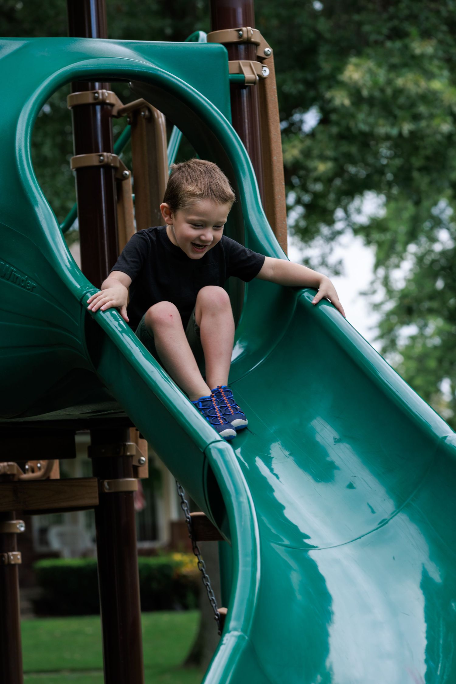 Young boy smiling on a green playground slide at an outdoor play structure on a cloudy day.