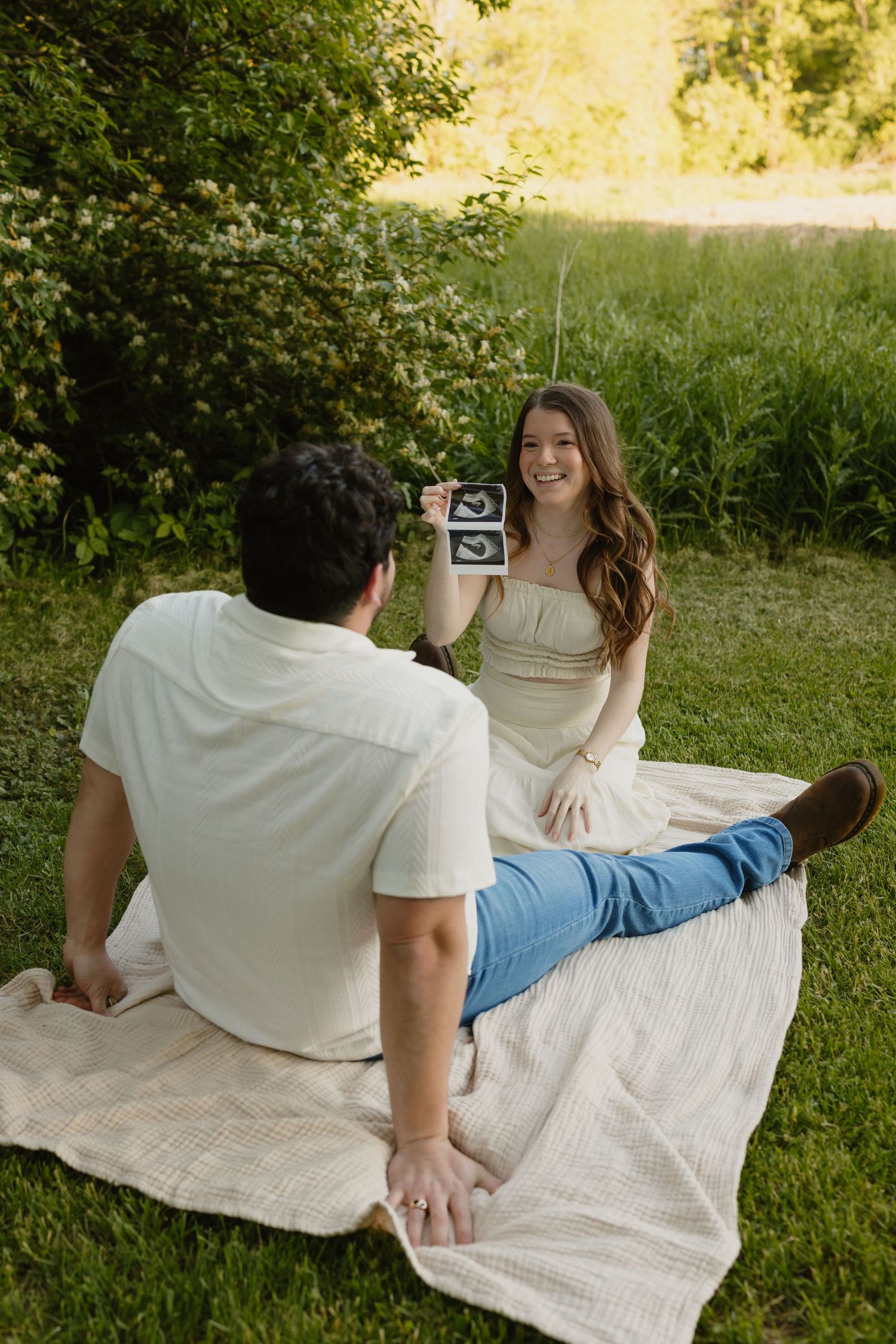 A smiling woman shows ultrasound photos to a man on a blanket in a sunny green park, announcing her pregnancy.