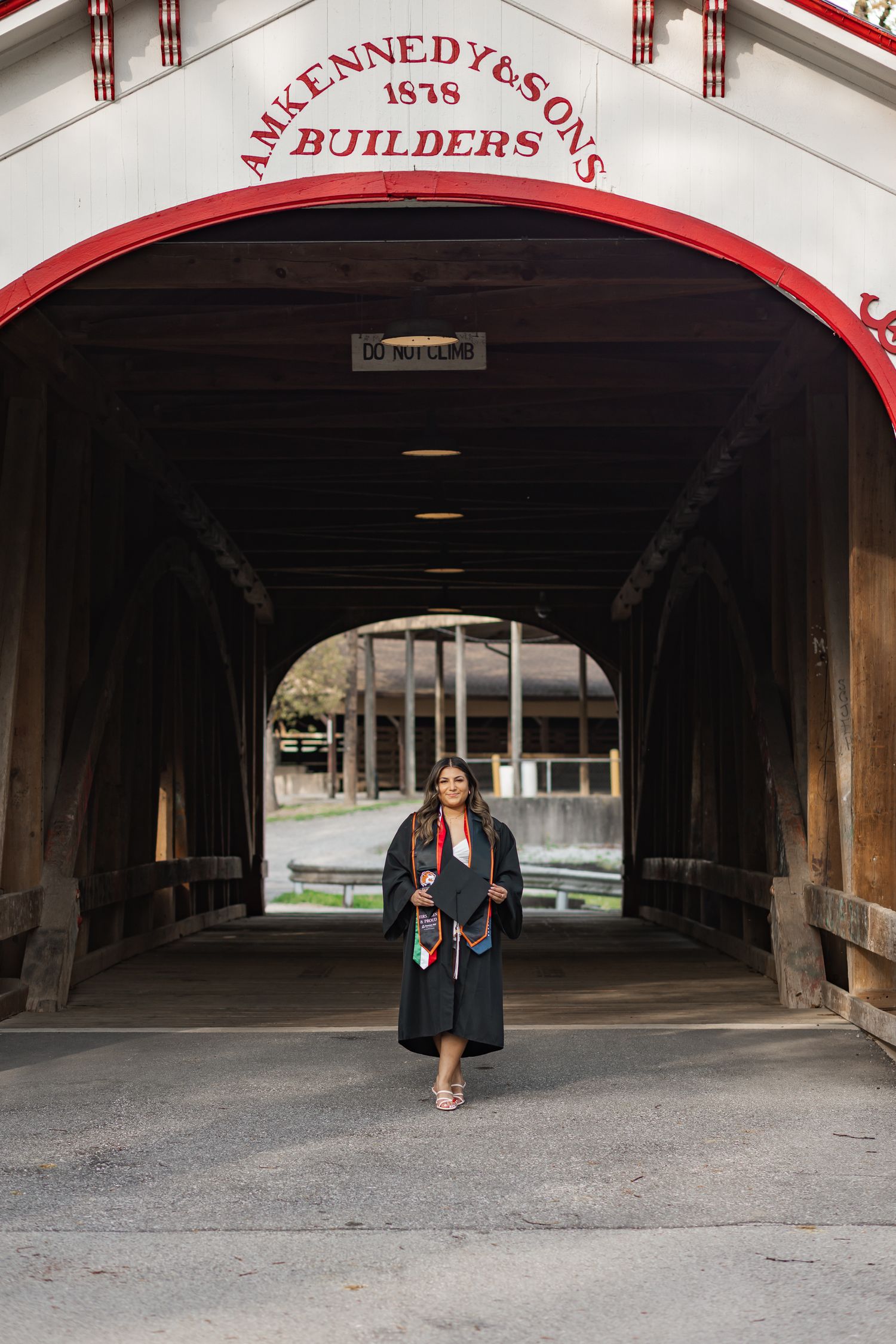 Graduate in cap and gown poses inside a historic covered bridge with AM Kennedy and Sons Builders 1878 signage.