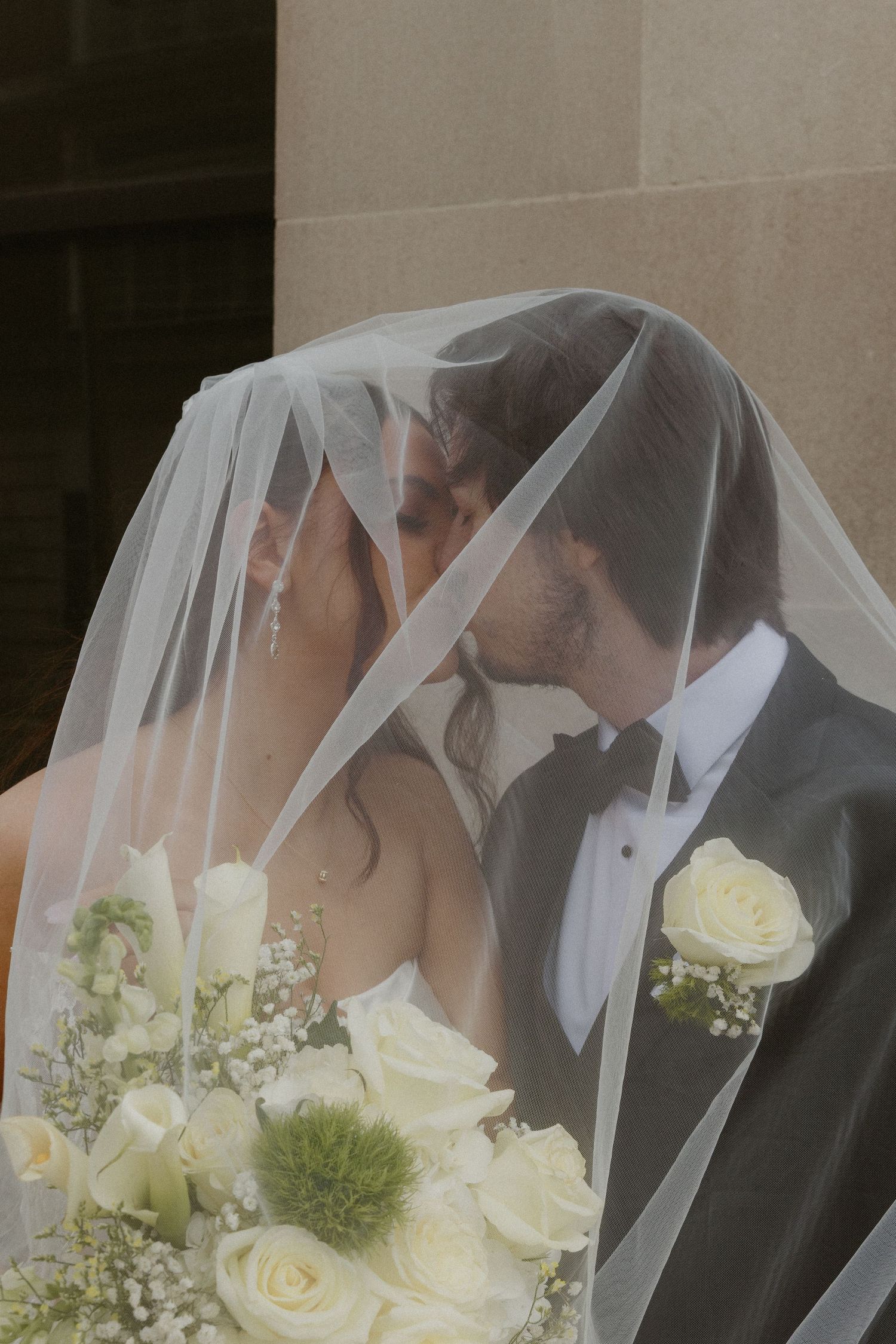Bride and groom sharing a romantic kiss under a white wedding veil, with a bouquet of white roses.