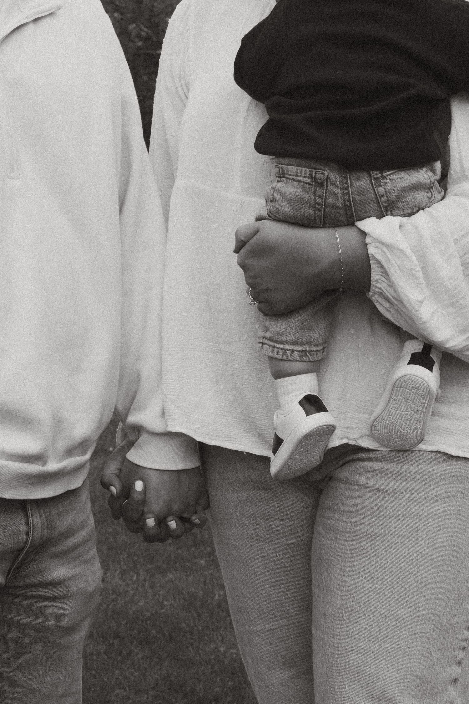 Black and white photo of a couple holding hands while carrying a baby in a denim jacket and white shoes.