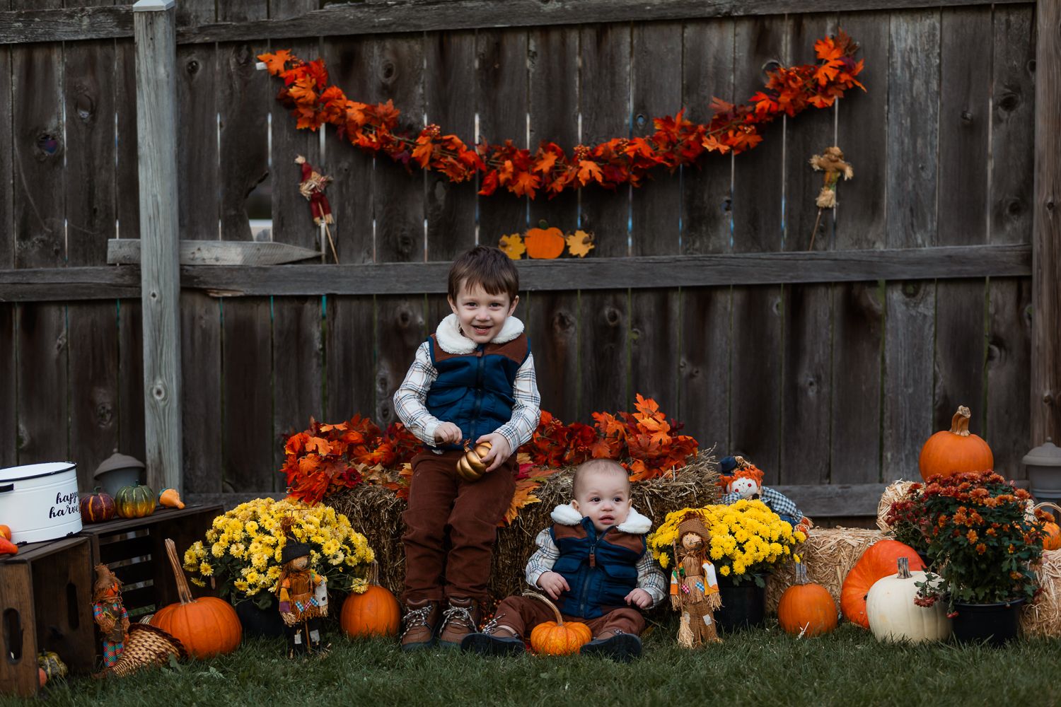 Two young children pose with fall pumpkins and flowers in front of a wooden fence decorated with autumn garlands.