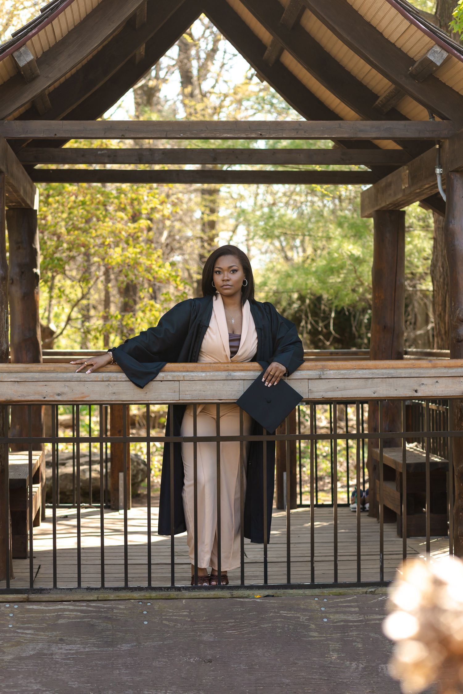 Graduate in black robe posing under wooden gazebo structure in autumn outdoor setting.