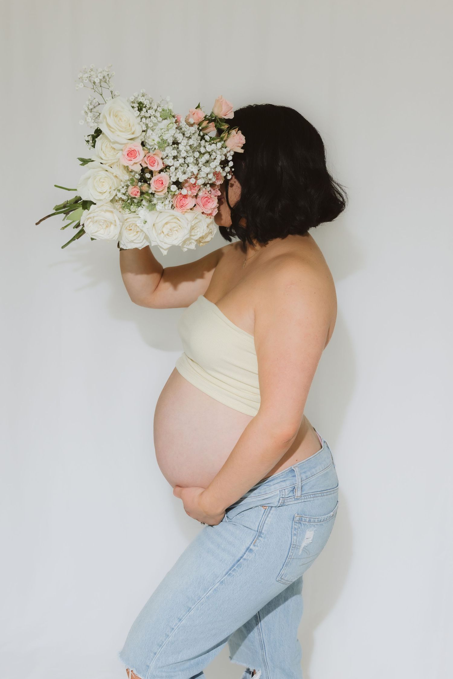 Pregnant woman in cream tube top and jeans holds bouquet of white and pink roses against white background.
